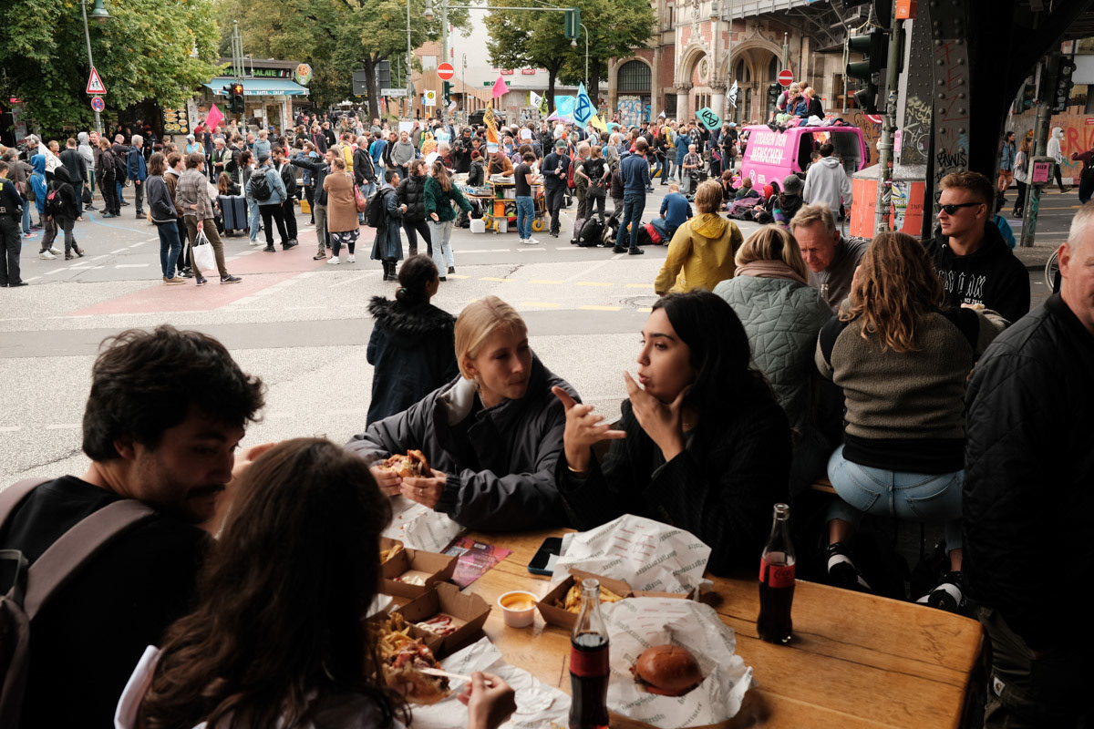 People eat burgers as activists block a major intersection in Berlin. Dozens of activists are glued or chained to the street, with hundreds of police at the scene.