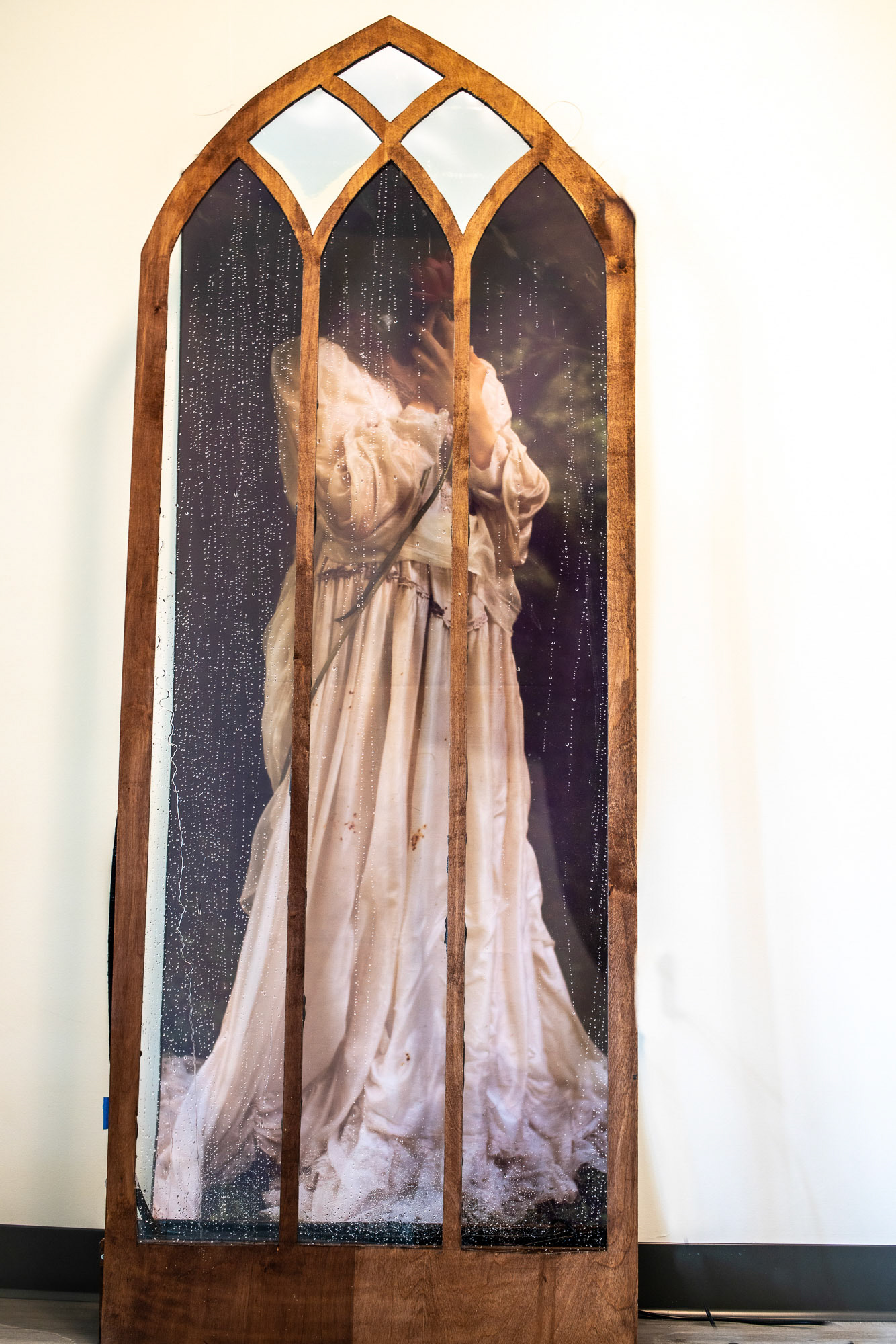 Forest Cathedral $10,500 70x30x24, (2023) Insulation art. A large canvas of a woman with a lilly hiding her face is framed by a church window edged in gold. The window has a fountain system that slowly rains water down the glass. The full effect is of looking out of a cathedral window through the rain to see one of the forest saints. standing among the trees.