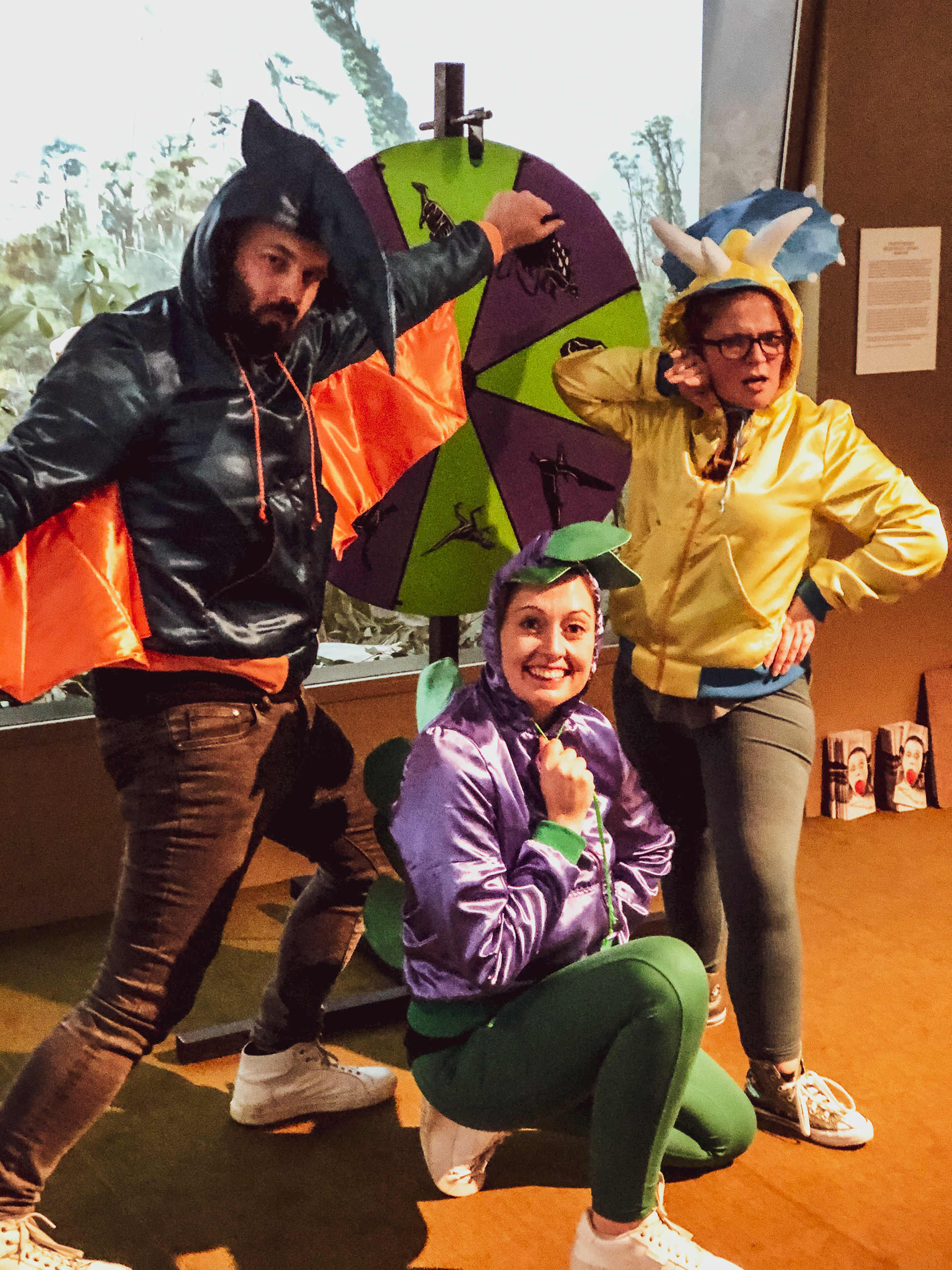 An image of the perfuct storm team posing in front of a wheel of fortune with dinosaurs on it. Dan is dressed as a Pterodactyl, Kathleen as a Stegosaurus, and Emma as a Triceratops.