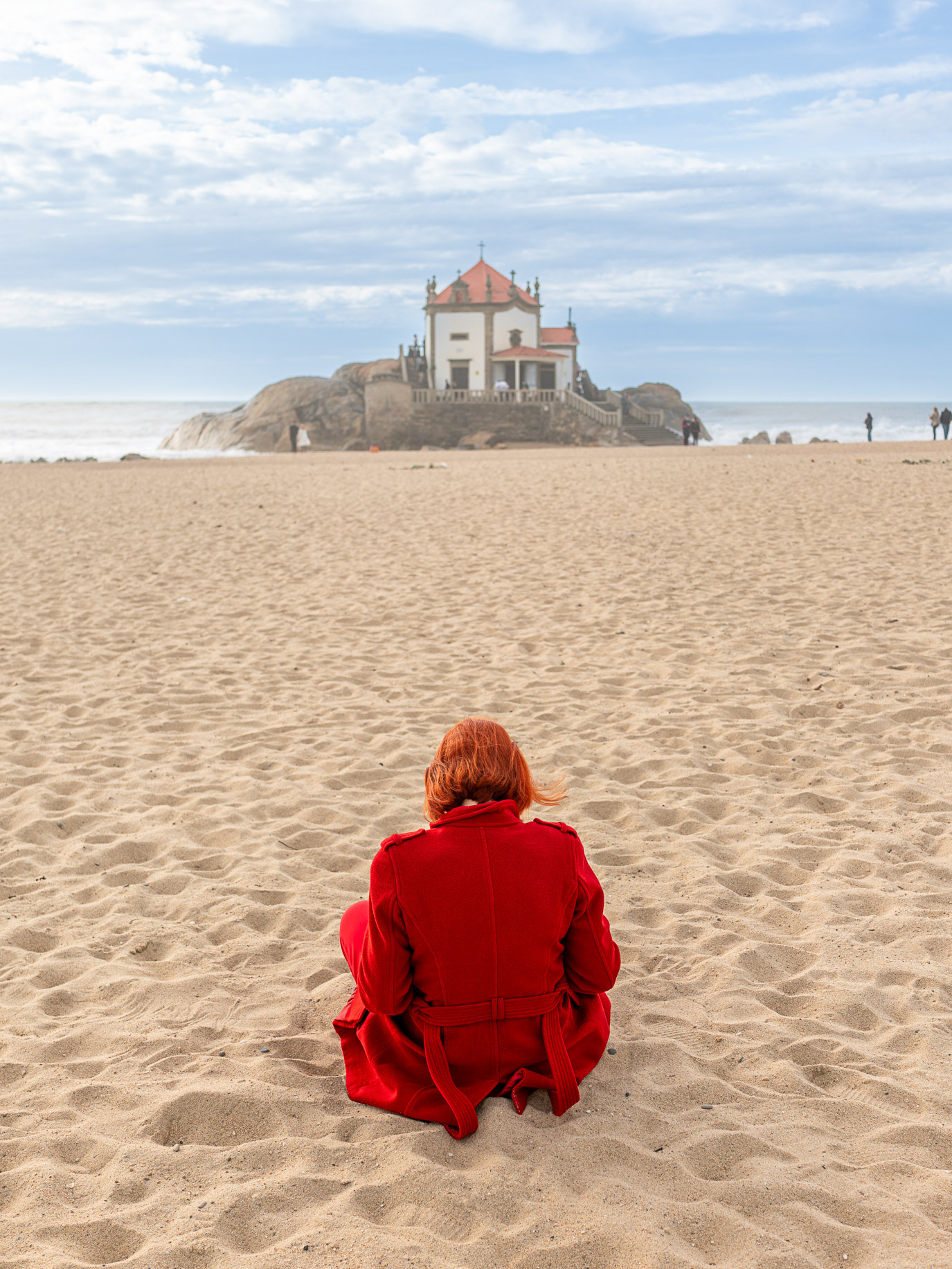 A woman in red seated on a beach facing a distant chapel in Vila Nova de Gaia, Portugal. Photograph by Alex Galmeanu.