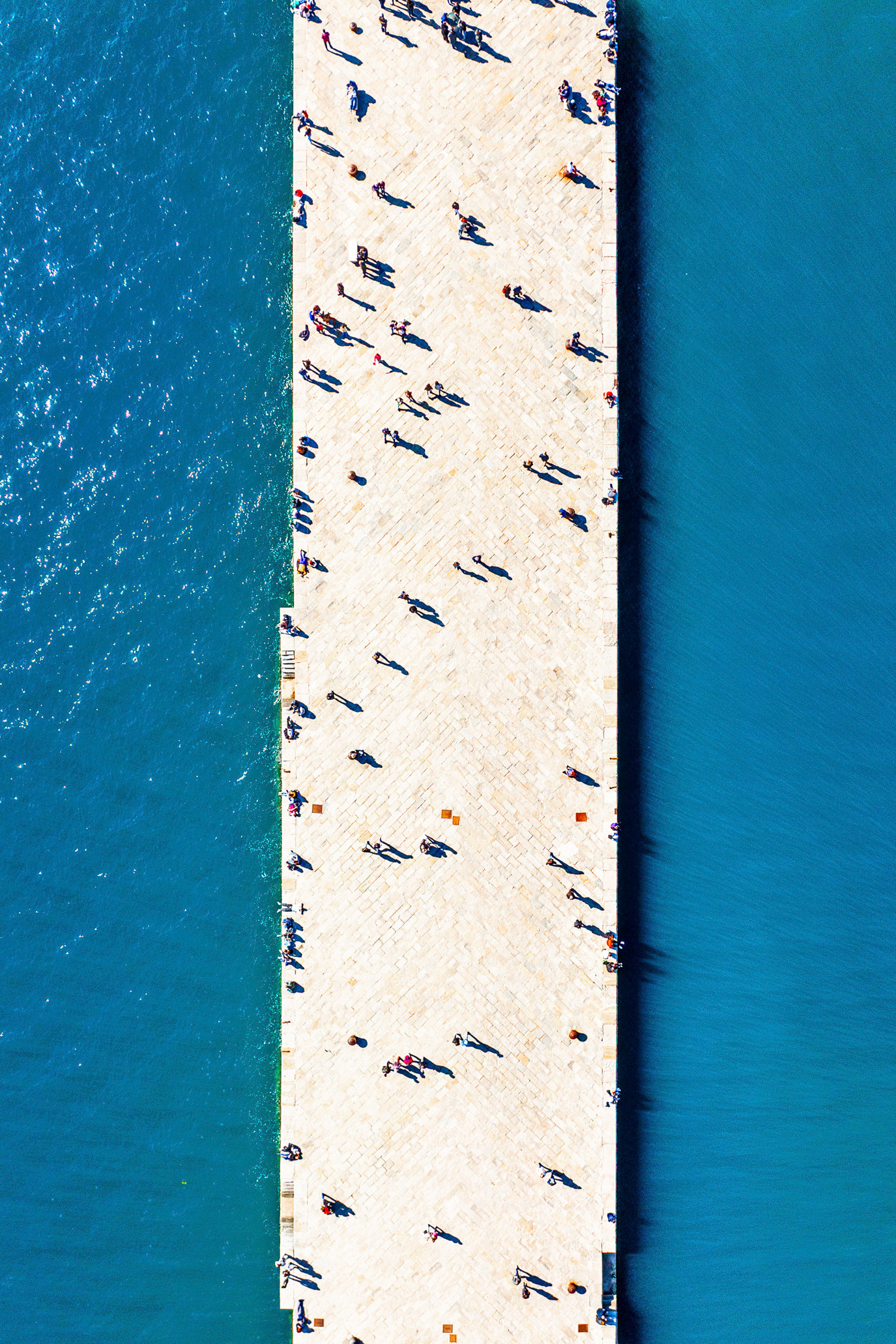 Tiny figures crossing a white pier between expanses of turquoise water in Trieste, Italy. Photograph by Alex Galmeanu.
