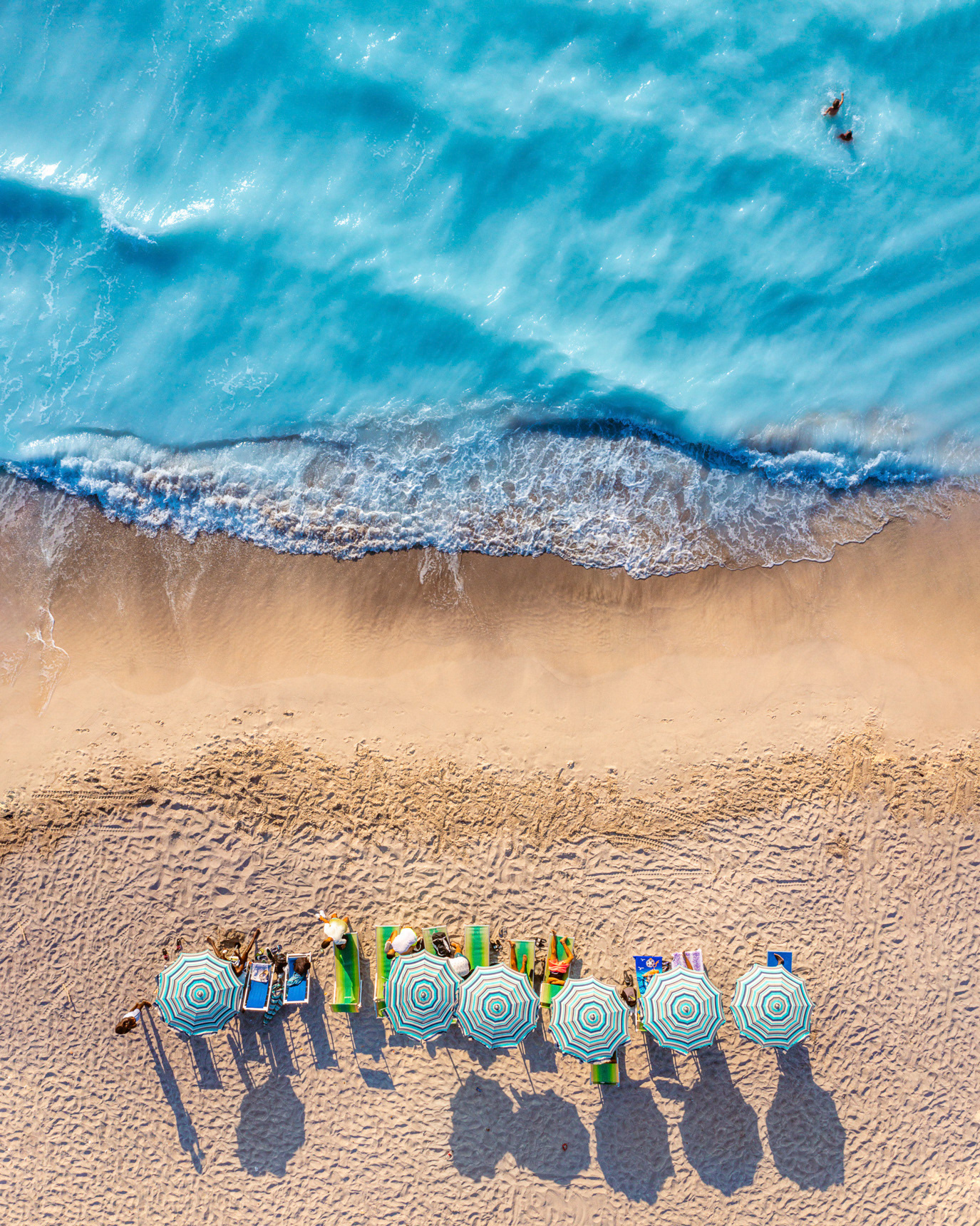 Beach umbrellas and long shadows beside turquoise water seen from above in Livorno, Italy. Photograph by Alex Galmeanu.