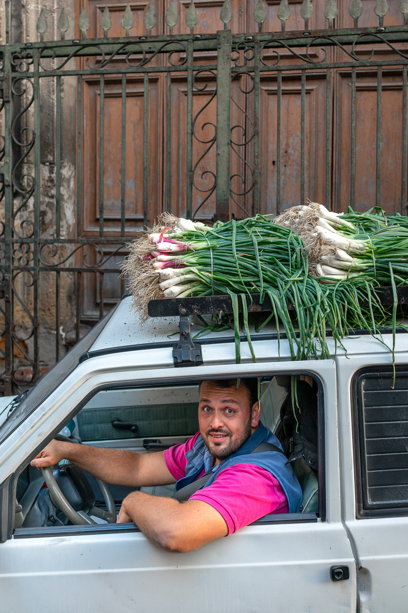 A smiling driver beneath bundles of fresh garlic tied to a small car in Palermo, Italy. Photograph by Alex Galmeanu.