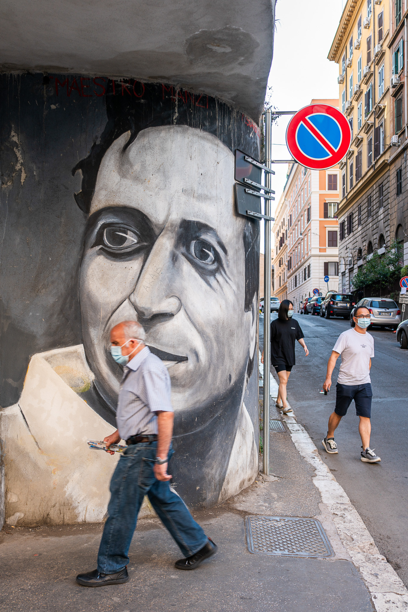 A masked pedestrian aligned with a monumental painted face in Rome, Italy. Photograph by Alex Galmeanu.