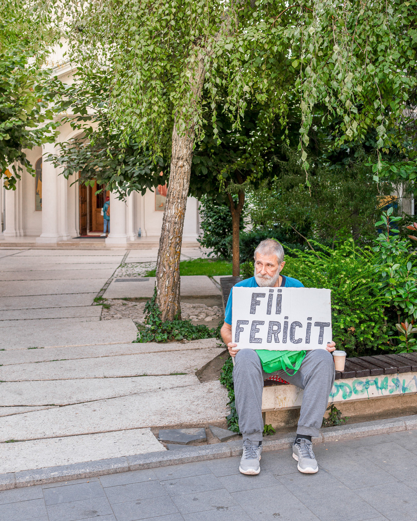 A seated man holding a handwritten “Fii fericit” sign in Bucharest, Romania. Photograph by Alex Galmeanu.