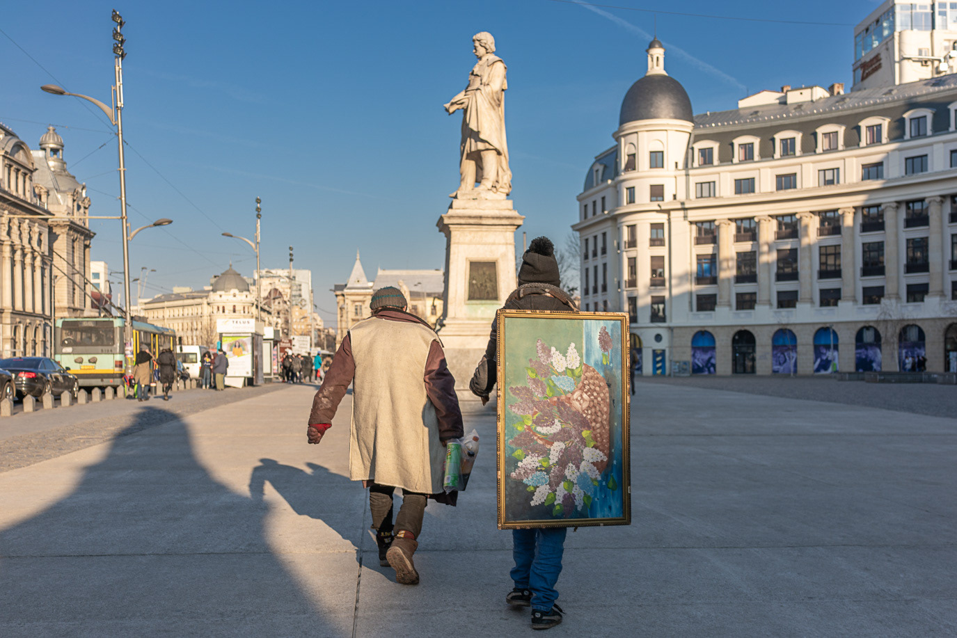 Two people carrying a flower painting across a public square in Bucharest, Romania. Photograph by Alex Galmeanu.