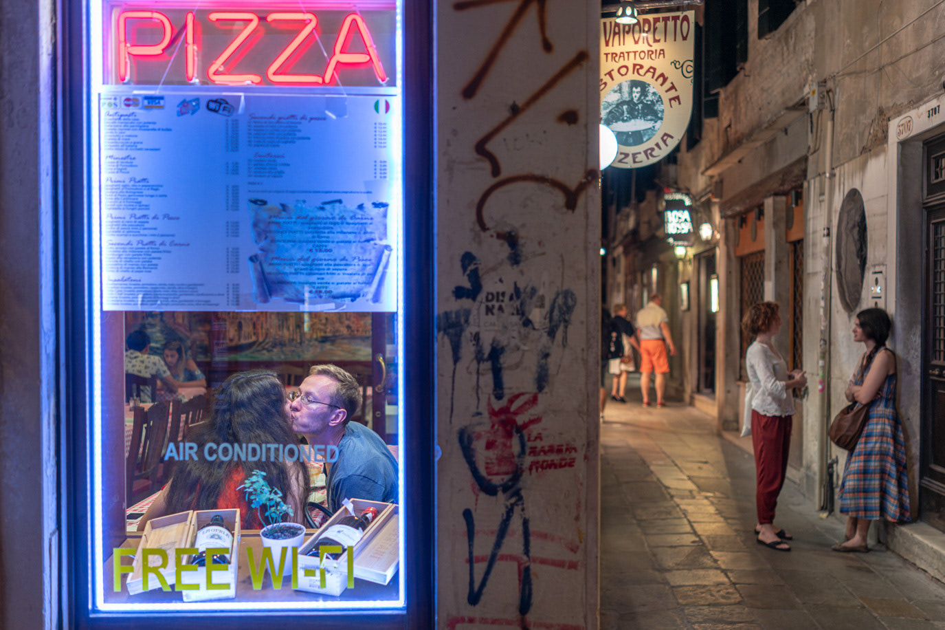 A seated diner framed by a neon pizza sign beside a narrow alley in Venice, Italy. Photograph by Alex Galmeanu.