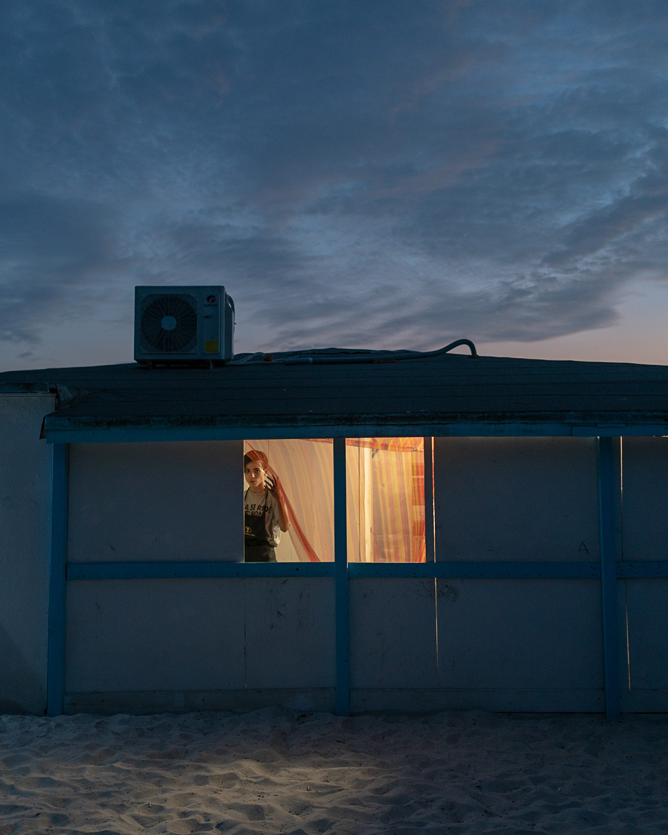 A lone figure framed in an illuminated beach cabin at dusk in Alghero, Italy. Photograph by Alex Galmeanu.