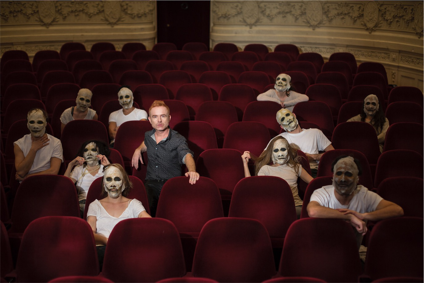 Radu Afrim seated in a theatre among figures wearing theatrical masks, photographed by Alex Gâlmeanu.
