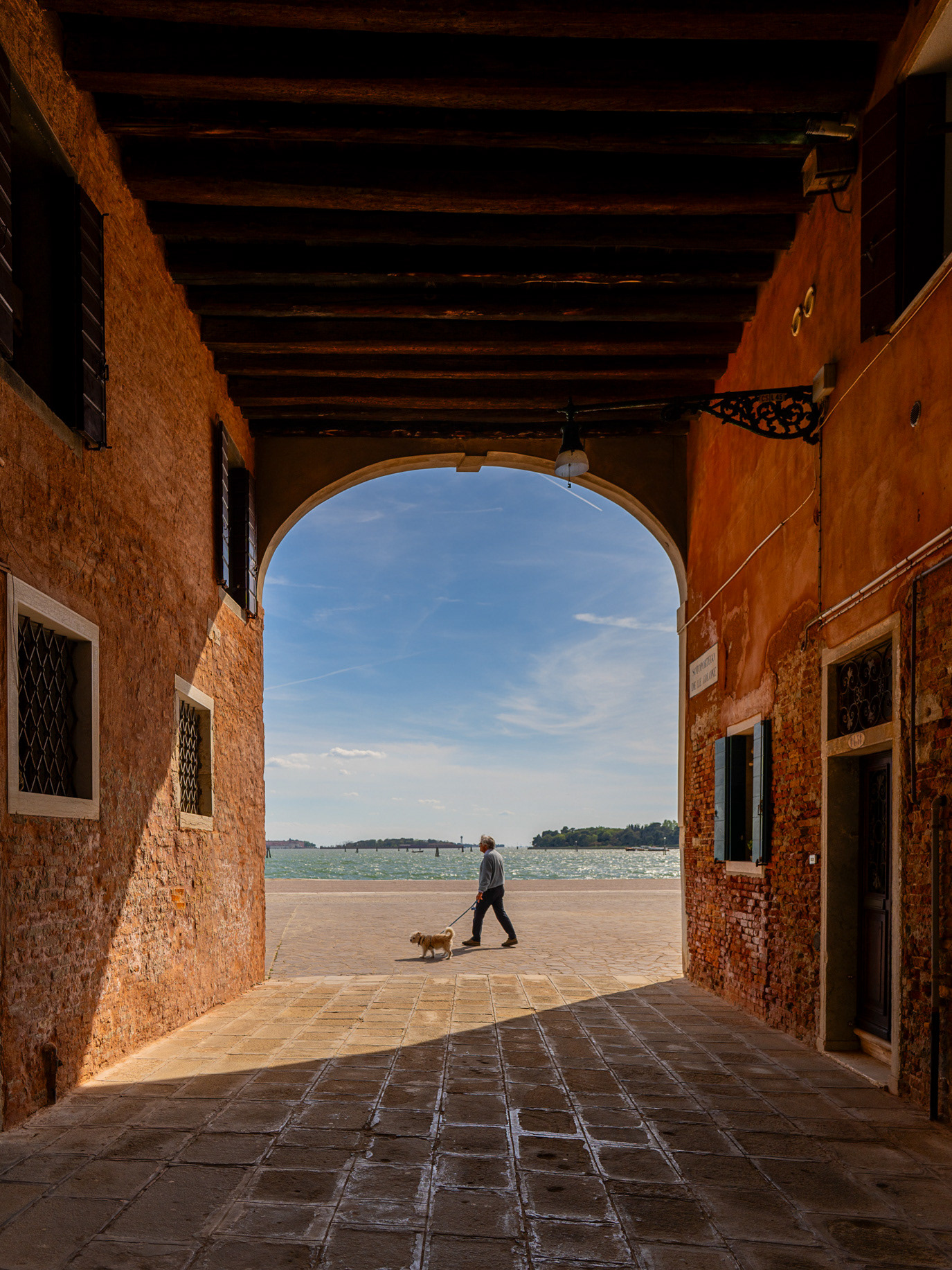 A solitary figure crossing a bright opening beneath a shadowed arch in Venice, Italy. Photograph by Alex Galmeanu.