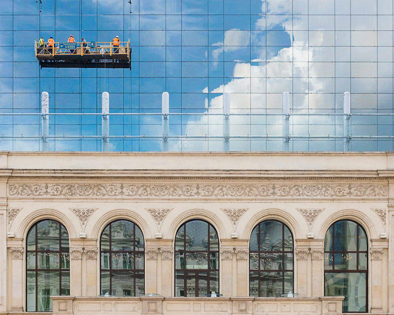 Window cleaners suspended against a glass facade filled with cloud reflections in Bucharest, Romania. Photograph by Alex Galmeanu.