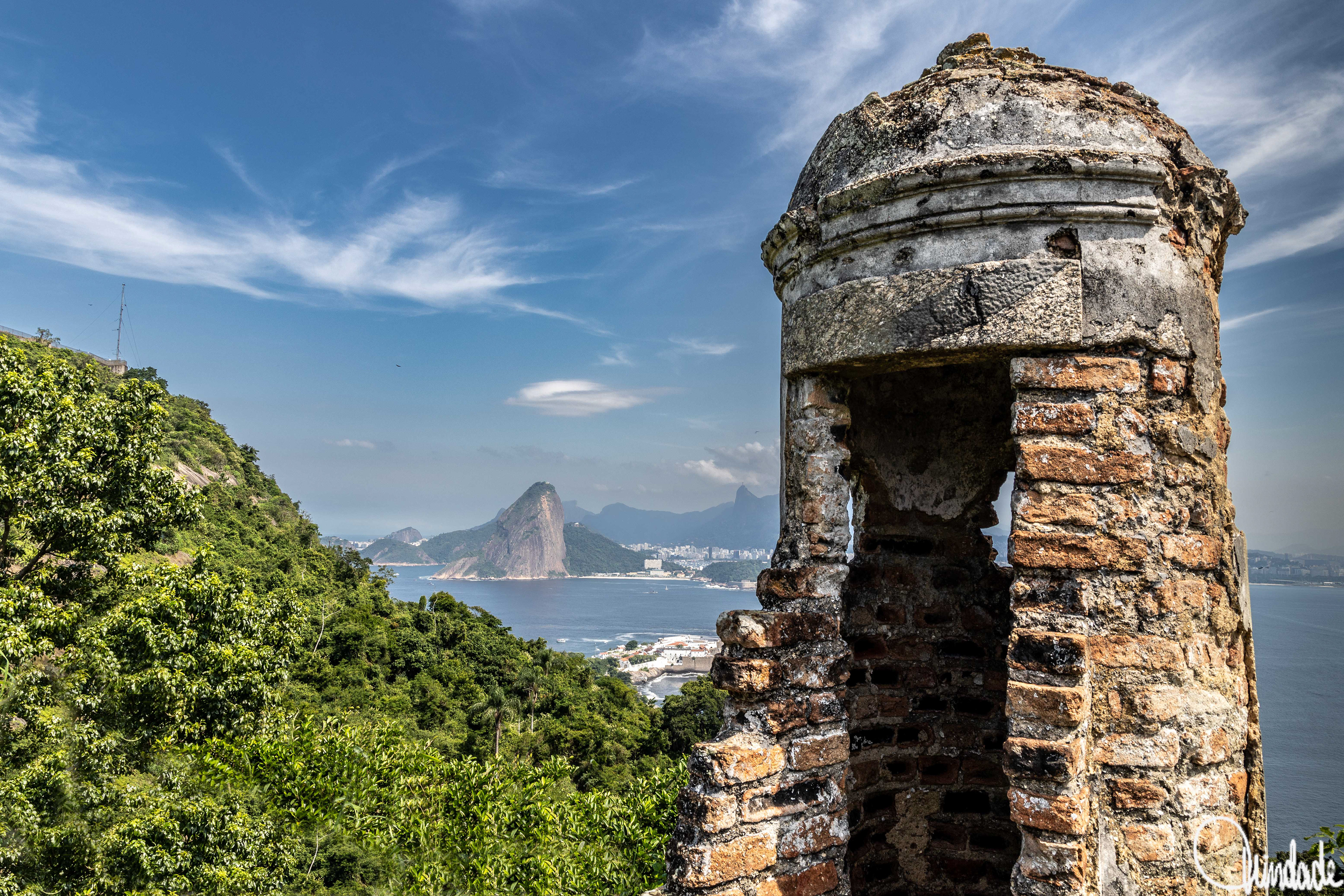 Guarita de pedra e tijolos envelhecidos com abertura em arco e cúpula no topo, em primeiro plano. Ao fundo, colinas verdes descem até o litoral e o Pão de Açúcar se destaca próximo ao mar sob céu azul com nuvens finas.