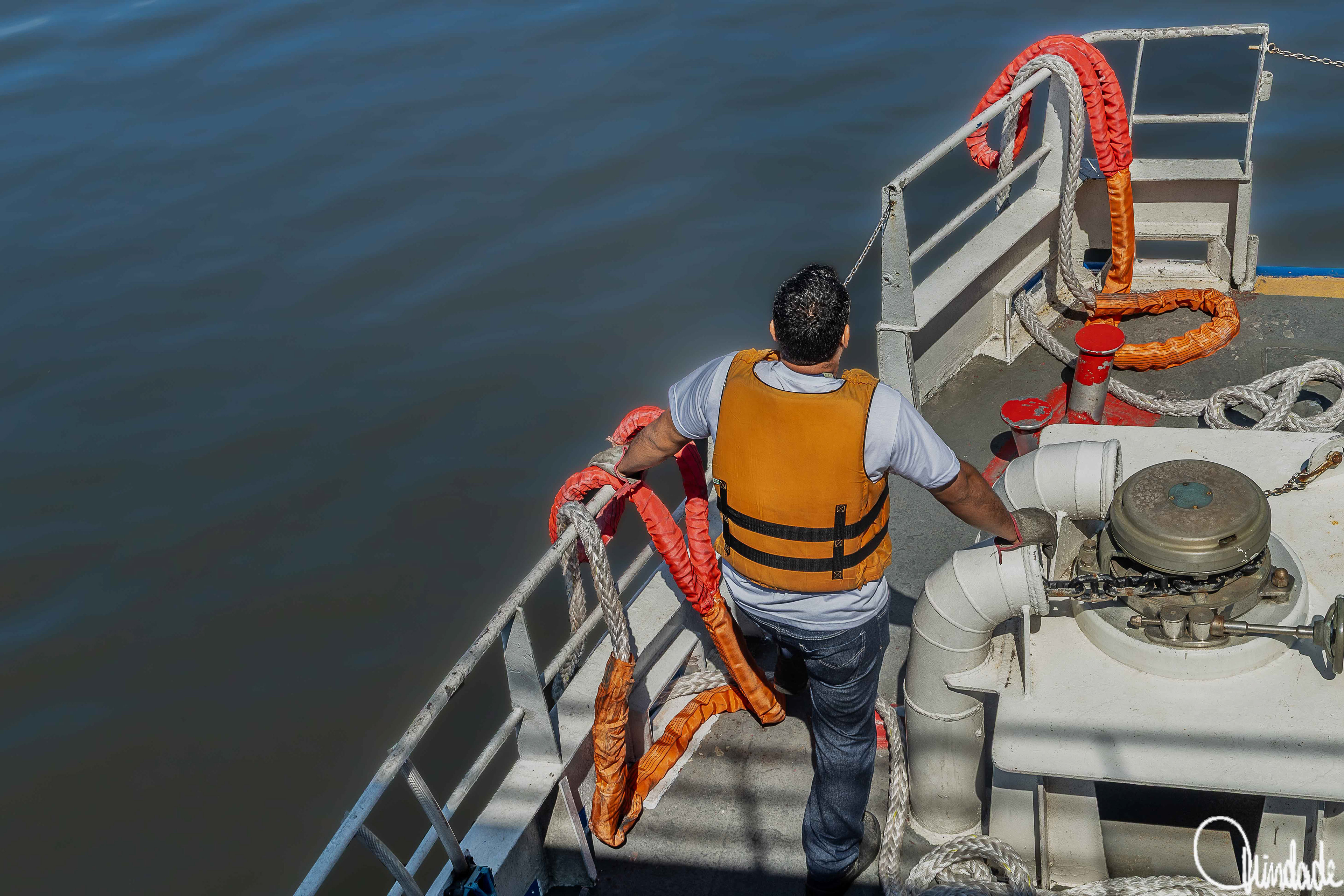 Pessoa de colete salva-vidas laranja em pé no convés de embarcação, segurando corda grossa com proteção laranja, com cabos enrolados ao redor e água calma ao fundo.