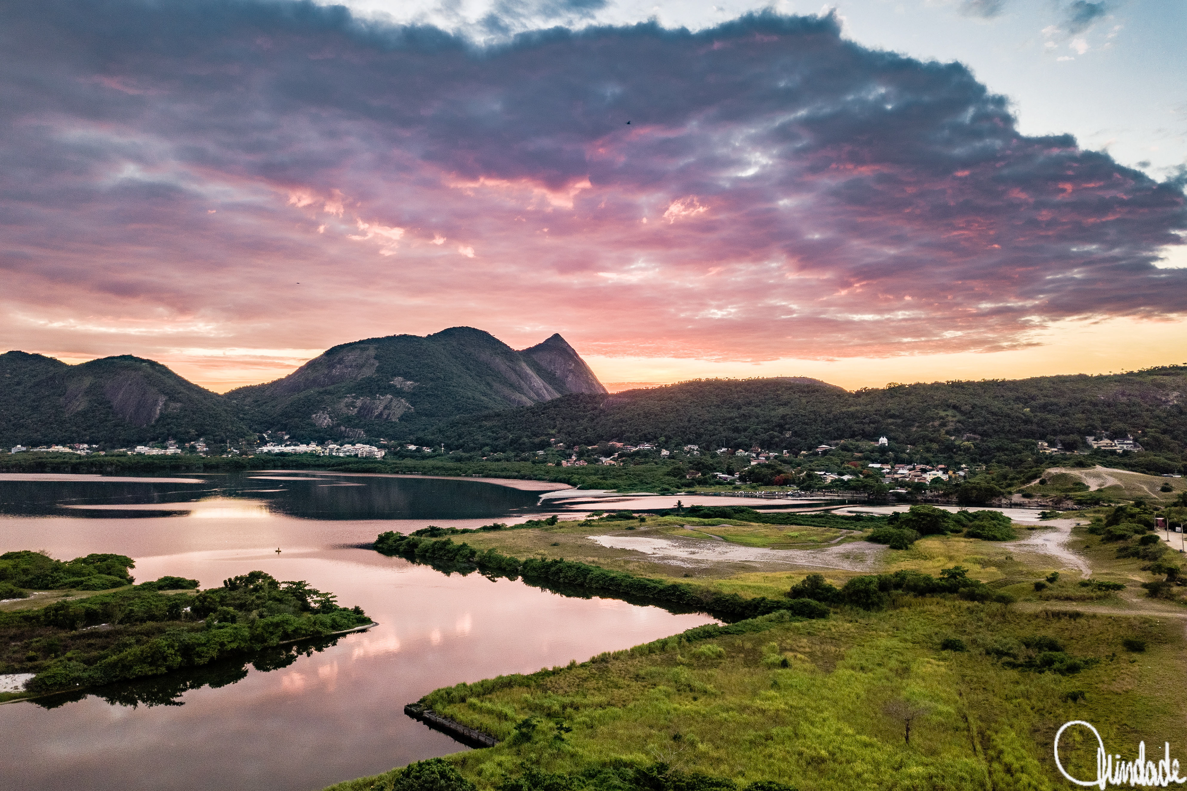 Entardecer na Laguna de Itaipu, Niterói, com águas calmas refletindo céu em tons de rosa e laranja. Serra da Tiririca coberta de vegetação verde ao fundo, pequenas casas na base das colinas completam a paisagem.
