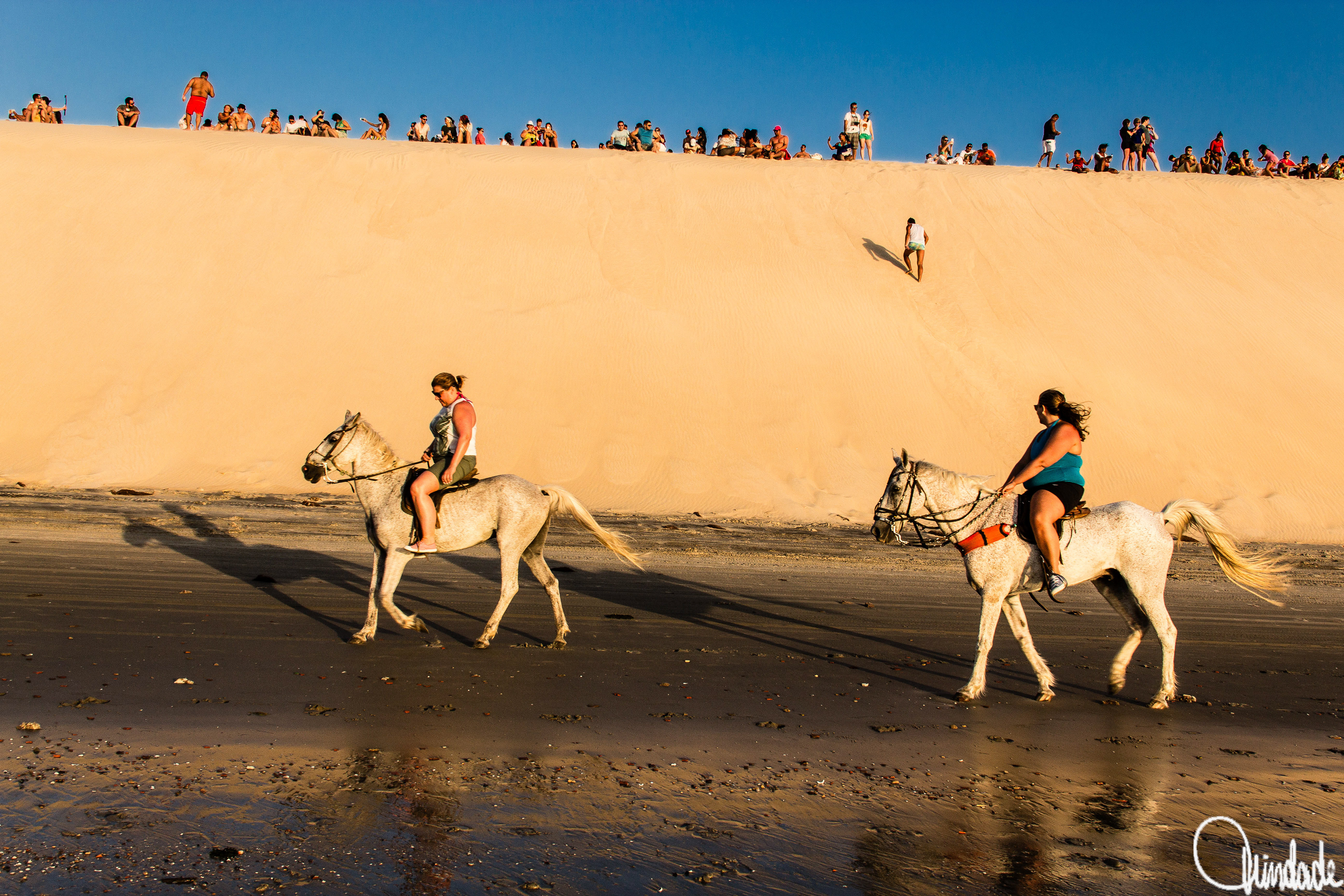 Turistas acomodados no alto de duna em Jericoacoara (CE) observam o pôr do sol, enquanto duas amazonas montam cavalos brancos na praia ao pé da duna.