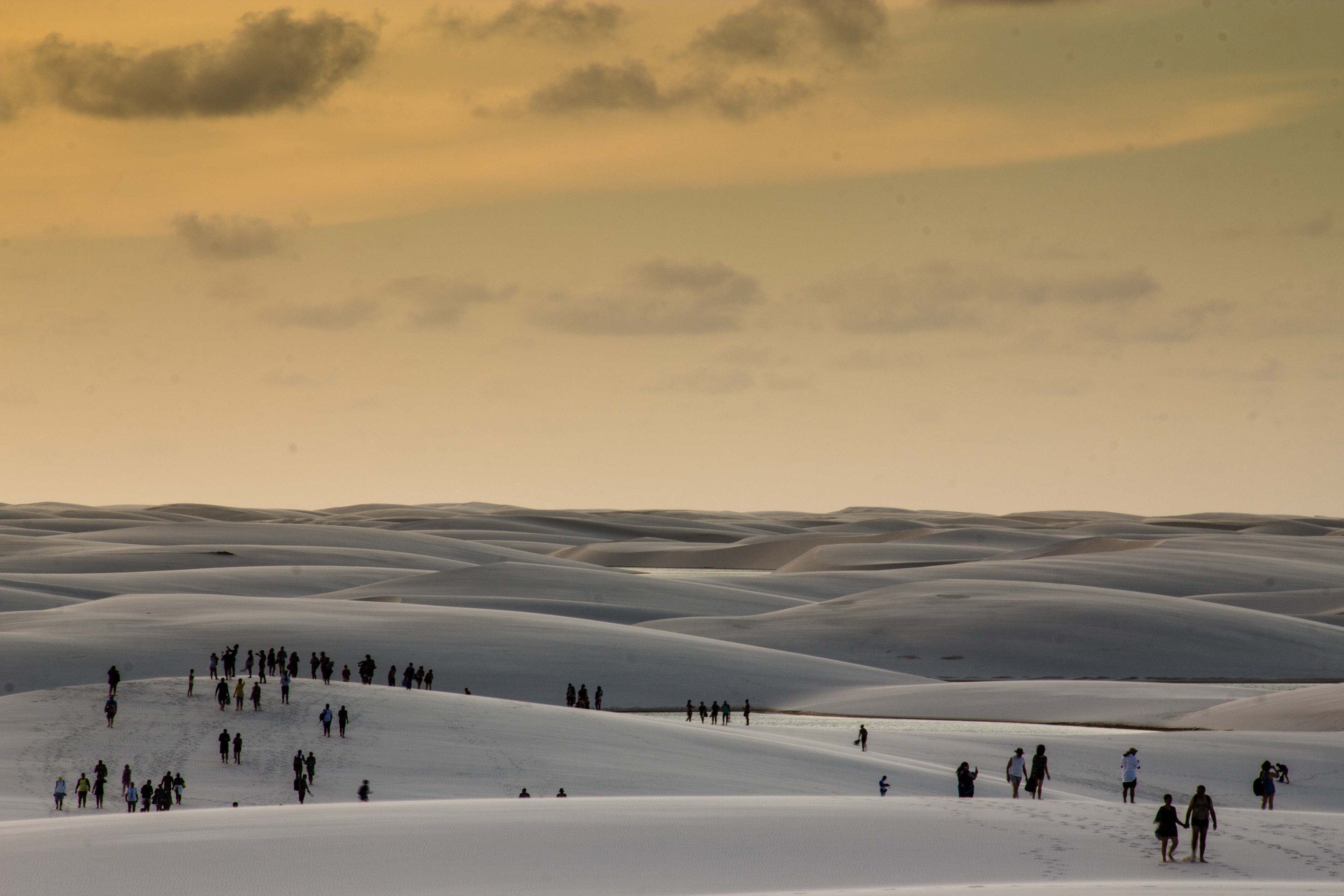    "Lençóis Maranhenses no Maranhão com dunas de areia branca sob céu dourado ao pôr do sol, grupos de pessoas caminhando e apreciando a paisagem, ressaltando a escala e beleza do cenário natural.