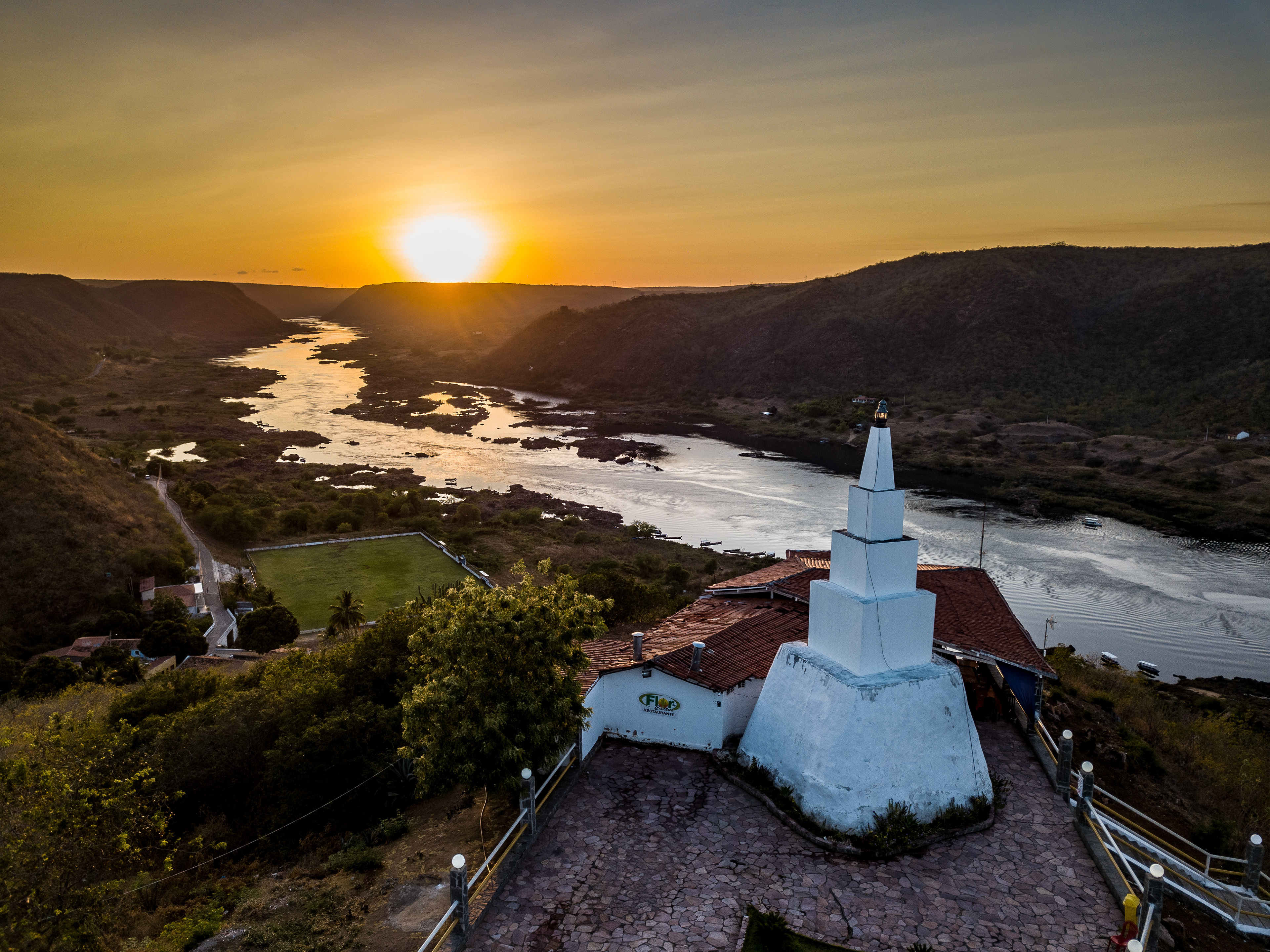 Mirante Secular em Piranhas, Sergipe, obelisco branco no alto de escadaria com vista panorâmica para o rio São Francisco ao pôr do sol, colinas ao redor e construções históricas próximas.