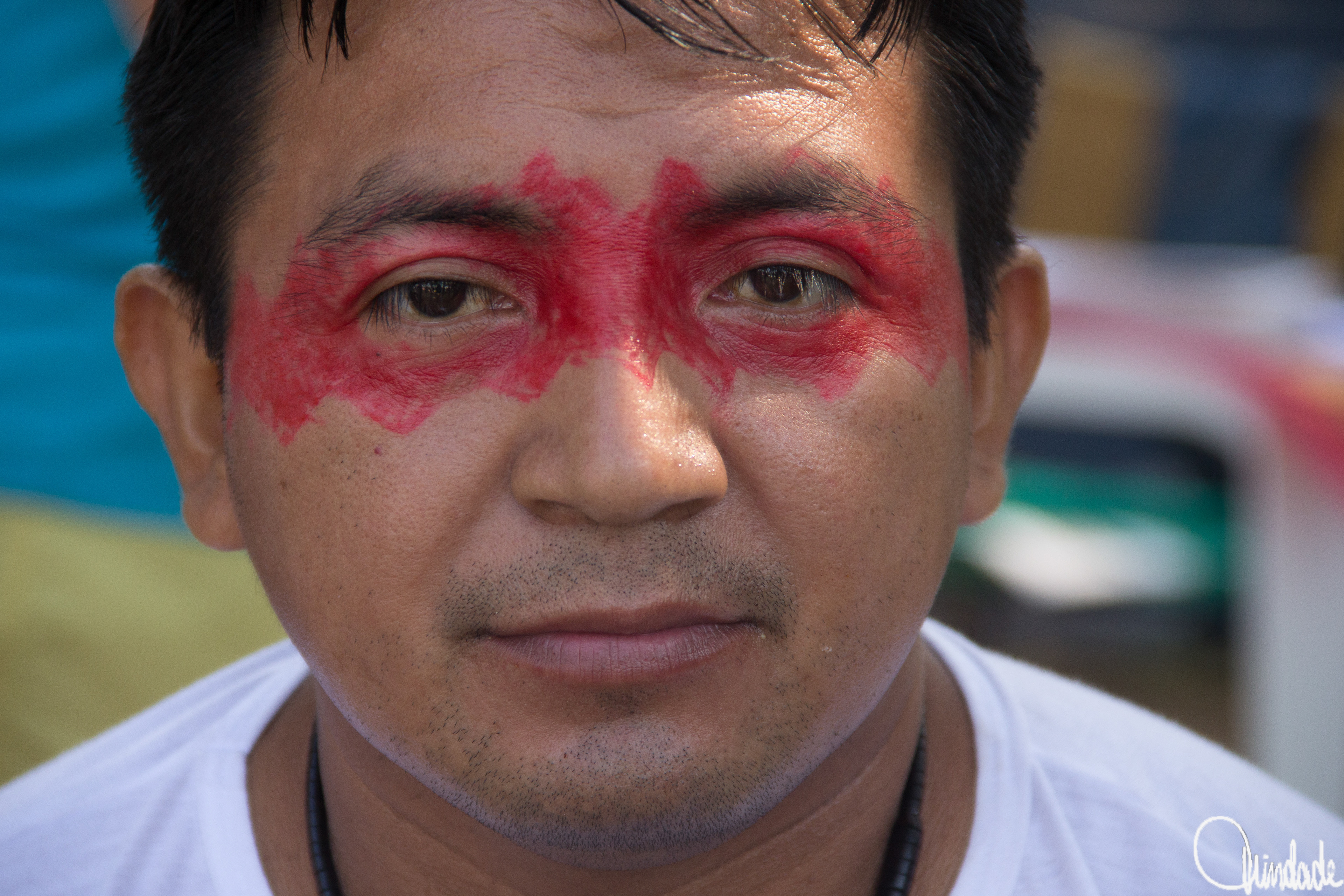 Retrato de homem com pintura vermelha simétrica no rosto, camisa branca e colar preto, em ambiente externo colorido, simbolizando tradição indígena sul-americana.