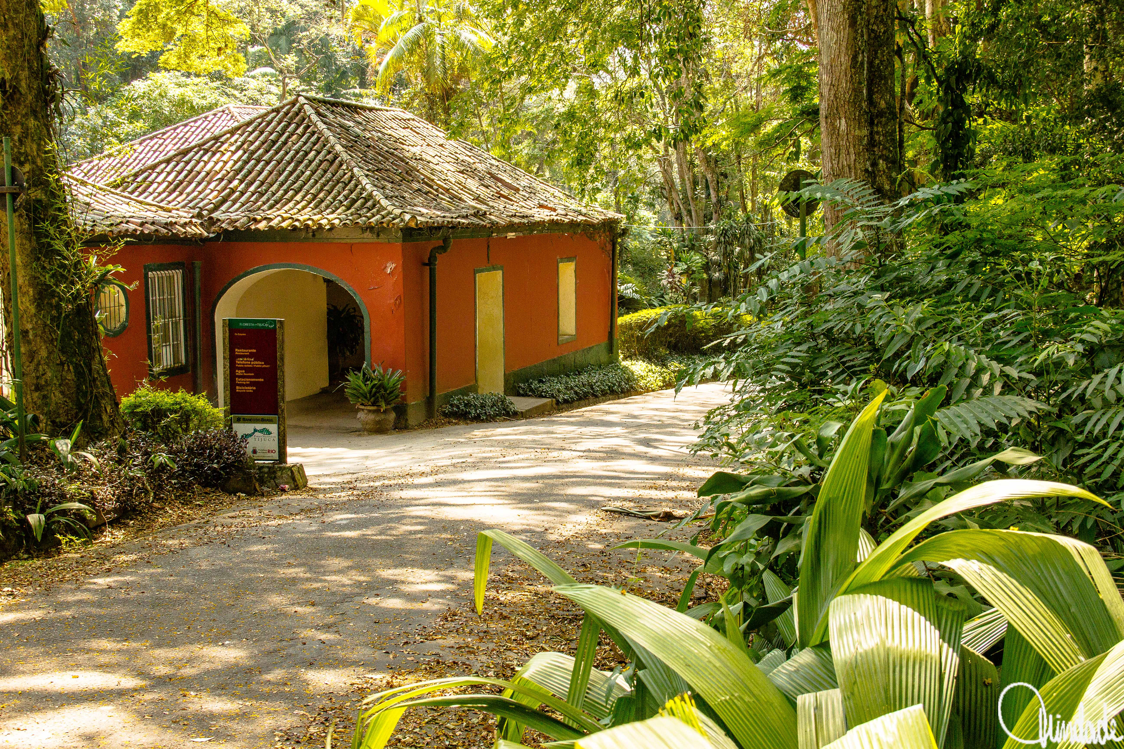 Construção rústica com paredes alaranjadas e telhado de barro, cercada por vegetação tropical. Entrada em arco com janelas gradeadas e plantas ornamentais. Estrutura histórica adaptada como restaurante dentro da Floresta da Tijuca