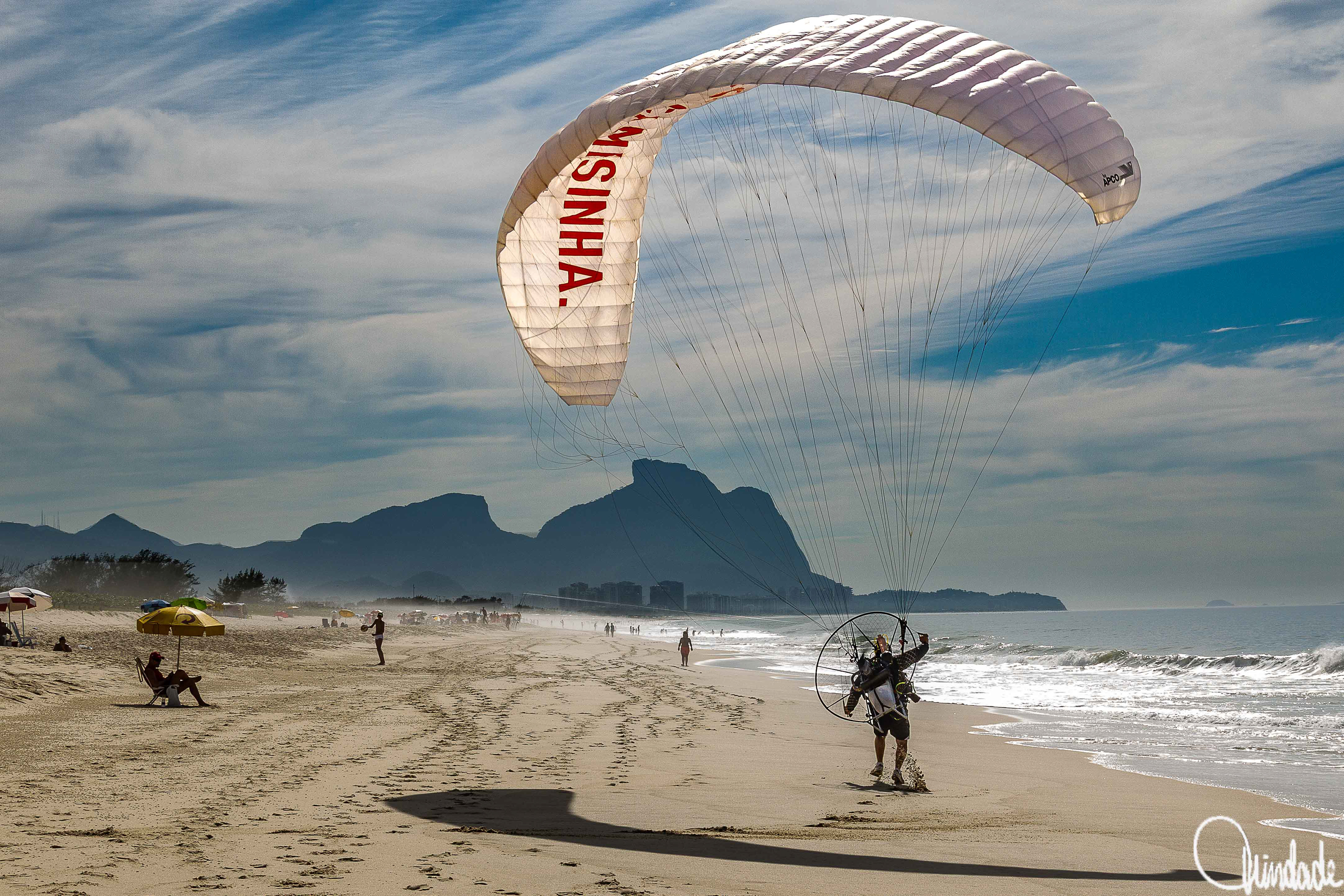 Pessoa praticando paramotor na areia da praia, com parapente branco com propaganda de uso de preservativo, mar ao fundo e montanhas ao horizonte (Praia da Reserva, Rio de Janeiro, RJ).