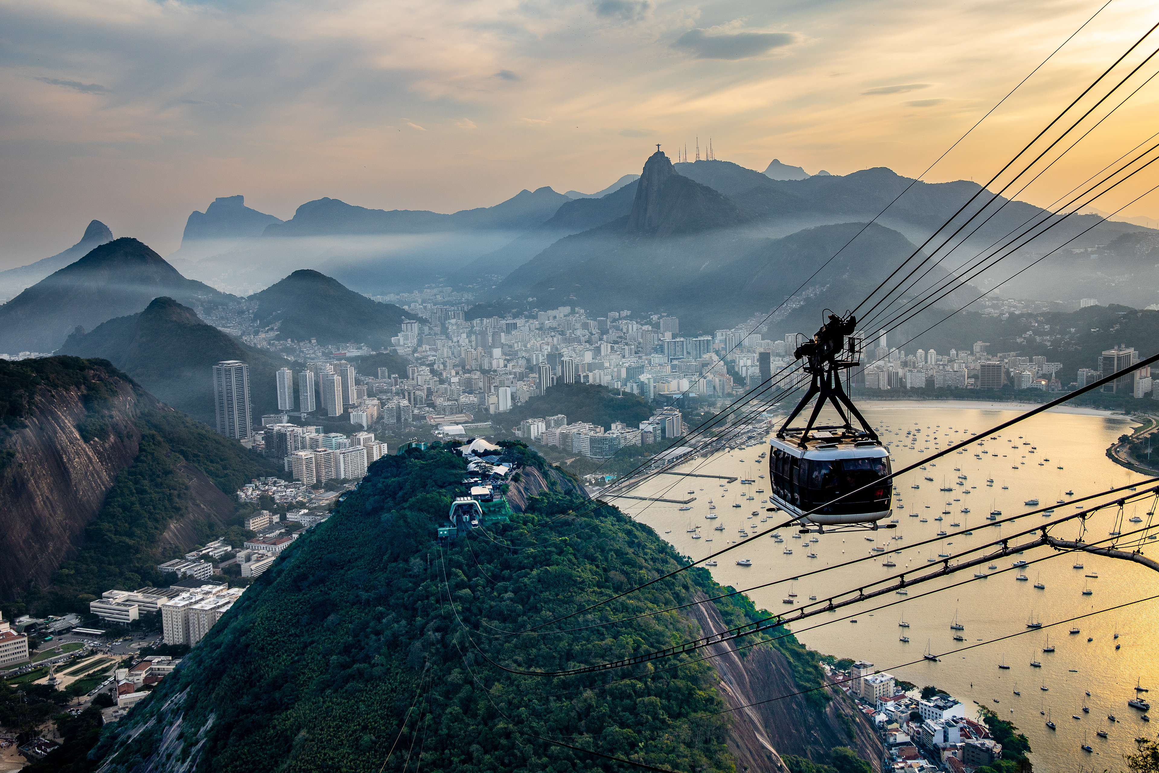    "Vista panorâmica da zona sul do Rio de Janeiro a partir do bondinho do Pão de Açúcar, com teleférico em primeiro plano, cidade e enseada com barcos iluminados pelo pôr do sol, Cristo Redentor visível ao fundo e maciço da Gávea destacado na paisagem.
