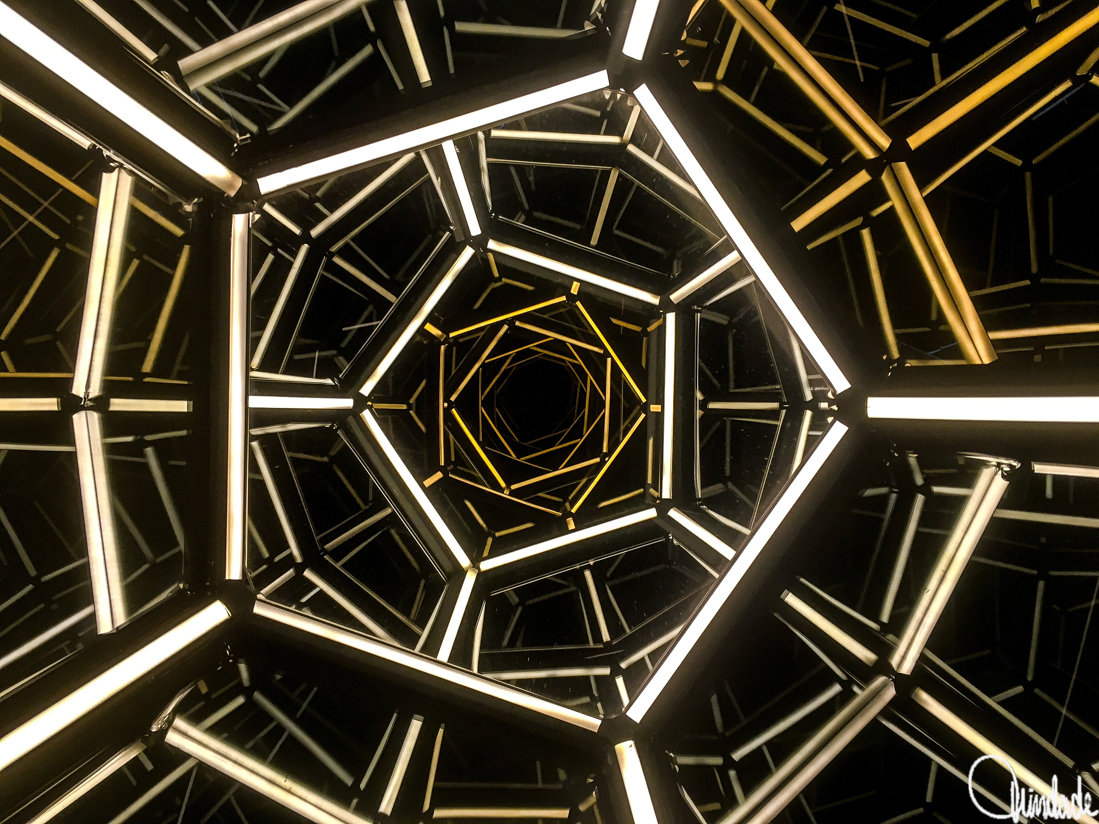 Túnel formado por estruturas geométricas pentagonais iluminadas em branco e amarelo. As luzes criam efeito de profundidade infinita e composição simétrica, lembrando um caleidoscópio.