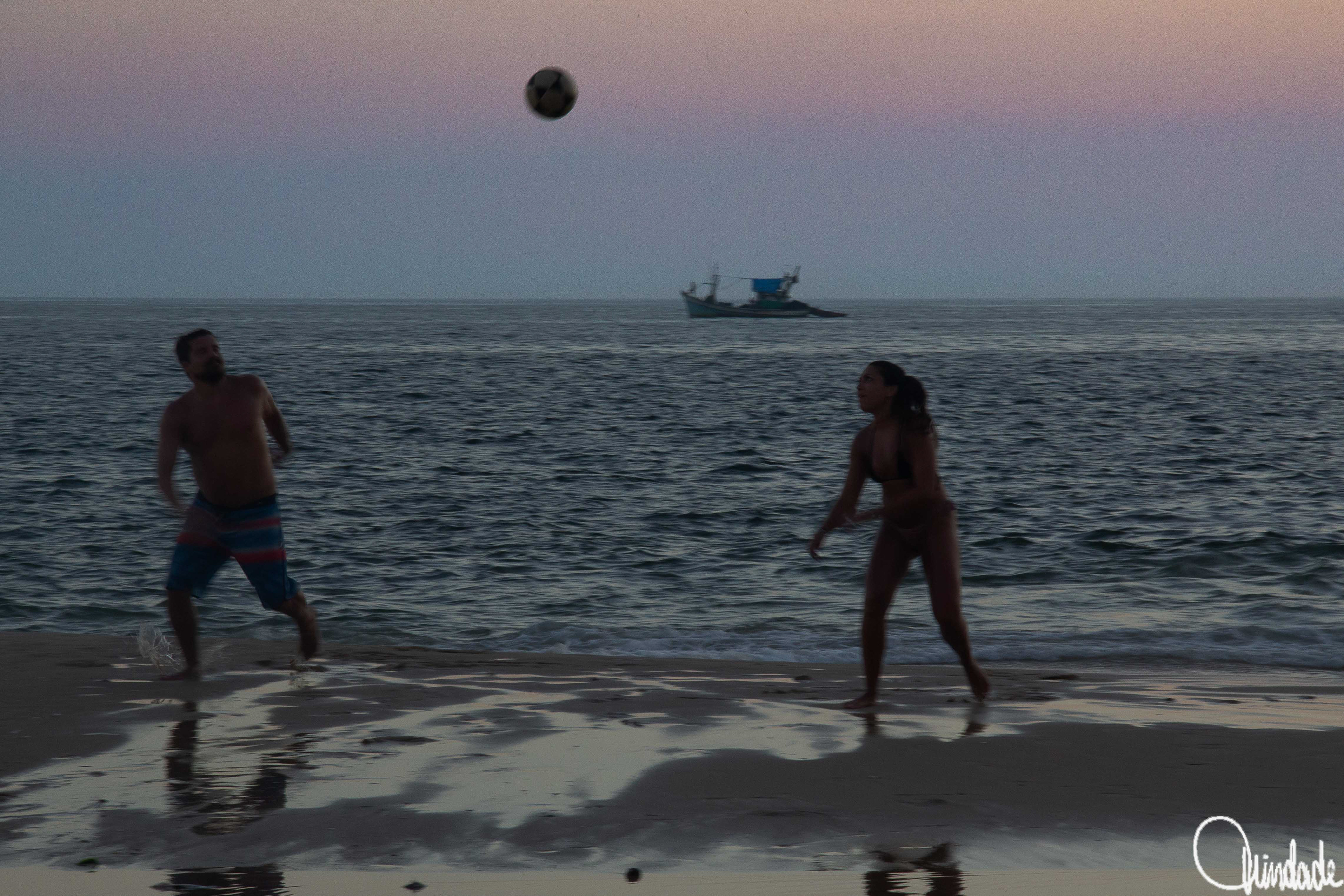 Dois jogadores de futevôlei em ação na areia da praia ao entardecer, com bola suspensa no ar, mar ao fundo e barco de pesca no horizonte sob céu em tons de rosa e azul.