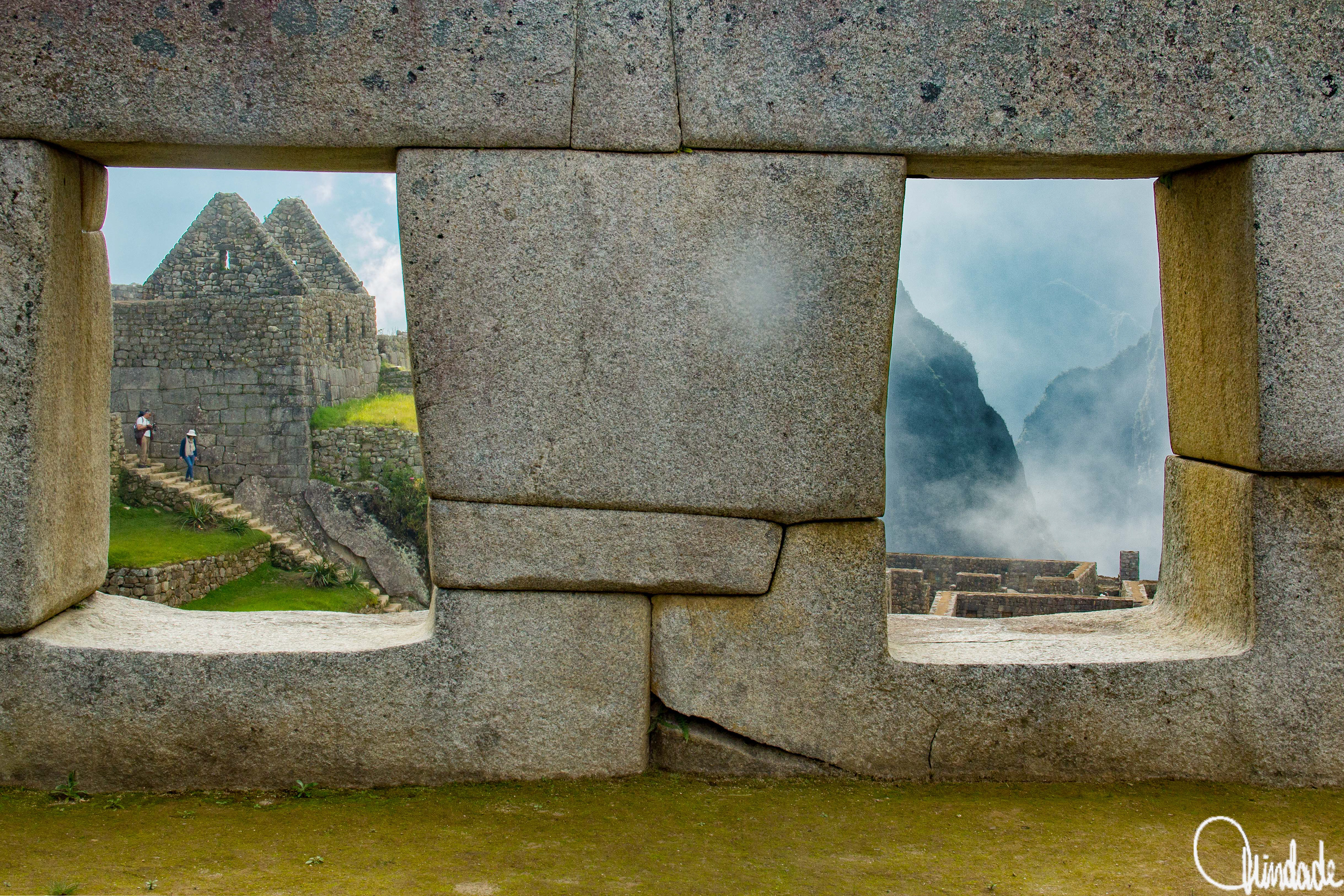 Parede de pedra inca com duas janelas retangulares visíveis, mostrando ao fundo estruturas de Machu Picchu e montanhas cobertas de névoa. Construção faz parte do Templo das Três Janelas, na Praça Sagrada da cidadela.
