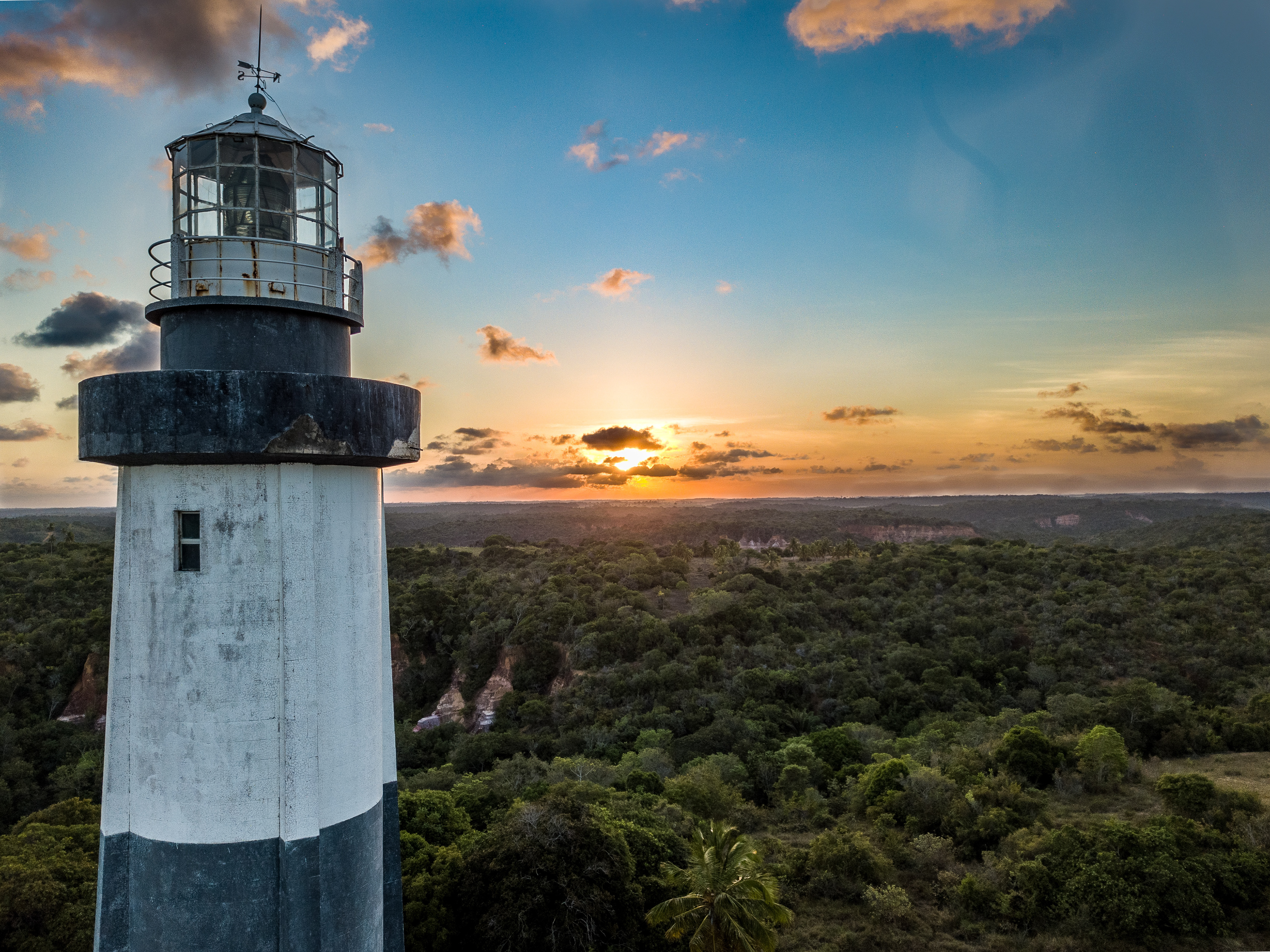    "Farol de Porto de Pedras em Alagoas, torre cilíndrica branca e cinza construída em 1933 pela Marinha, localizado no alto de colina com vista panorâmica para o mar e vegetação costeira ao pôr do sol.