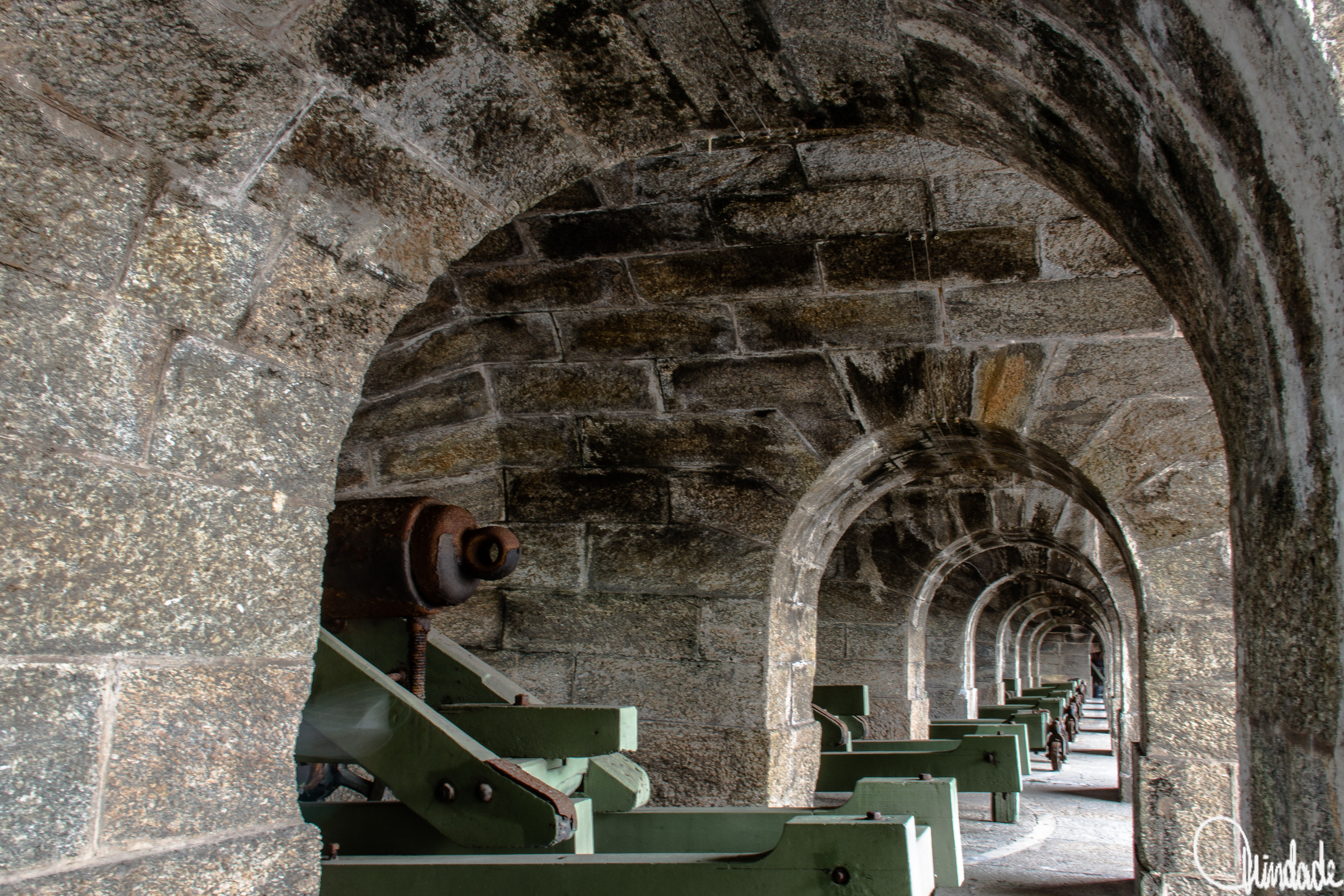 Corredor de fortificação com sucessivos arcos de pedra envelhecida, cada um abrigando suportes metálicos de canhões com rodas enferrujadas. A perspectiva destaca a repetição dos arcos e a simetria da arquitetura militar