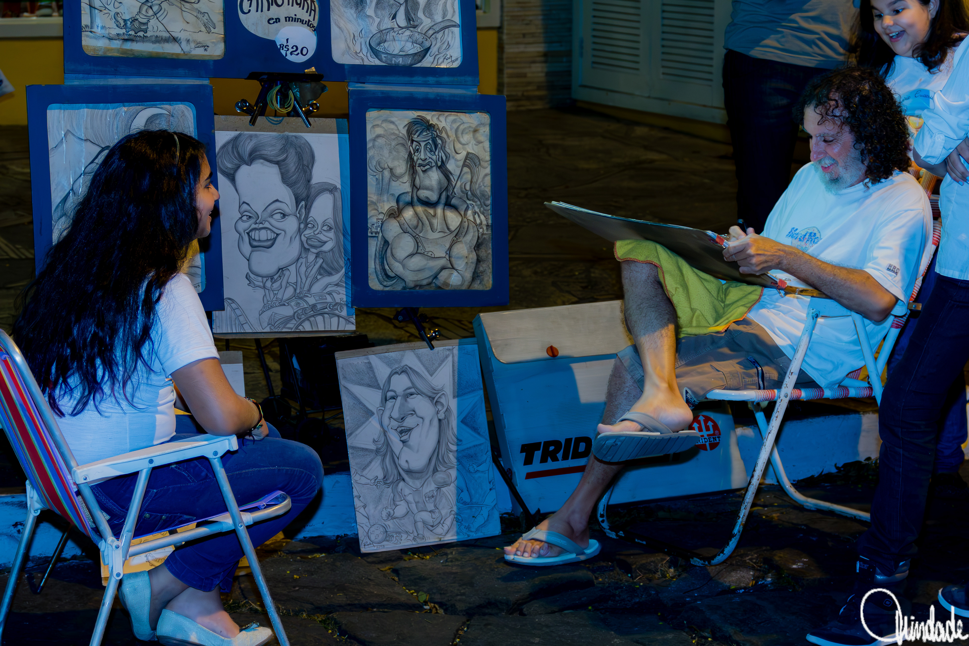 Artista de rua sentado desenha caricatura de um homem sorridente em feira urbana, com exemplos expostos e cartaz indicando preço de R$20.