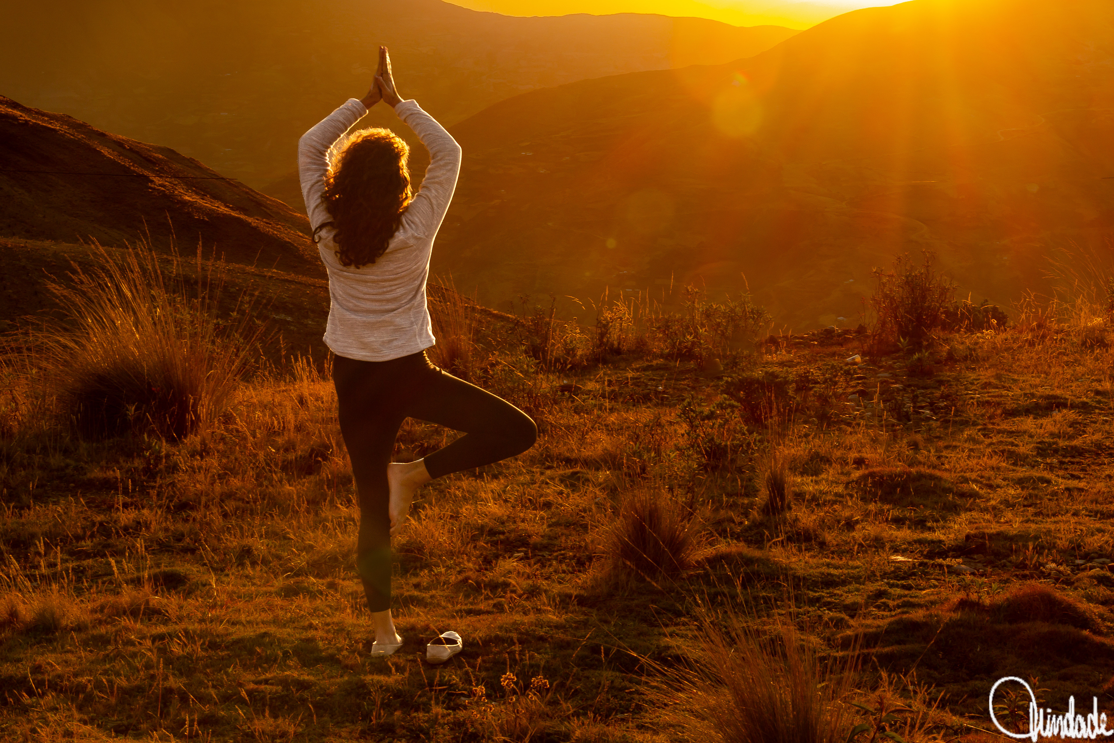 Pessoa em colina gramada faz pose de yoga ao pôr do sol na Cordilheira dos Andes, com luz dourada iluminando montanhas e vegetação.