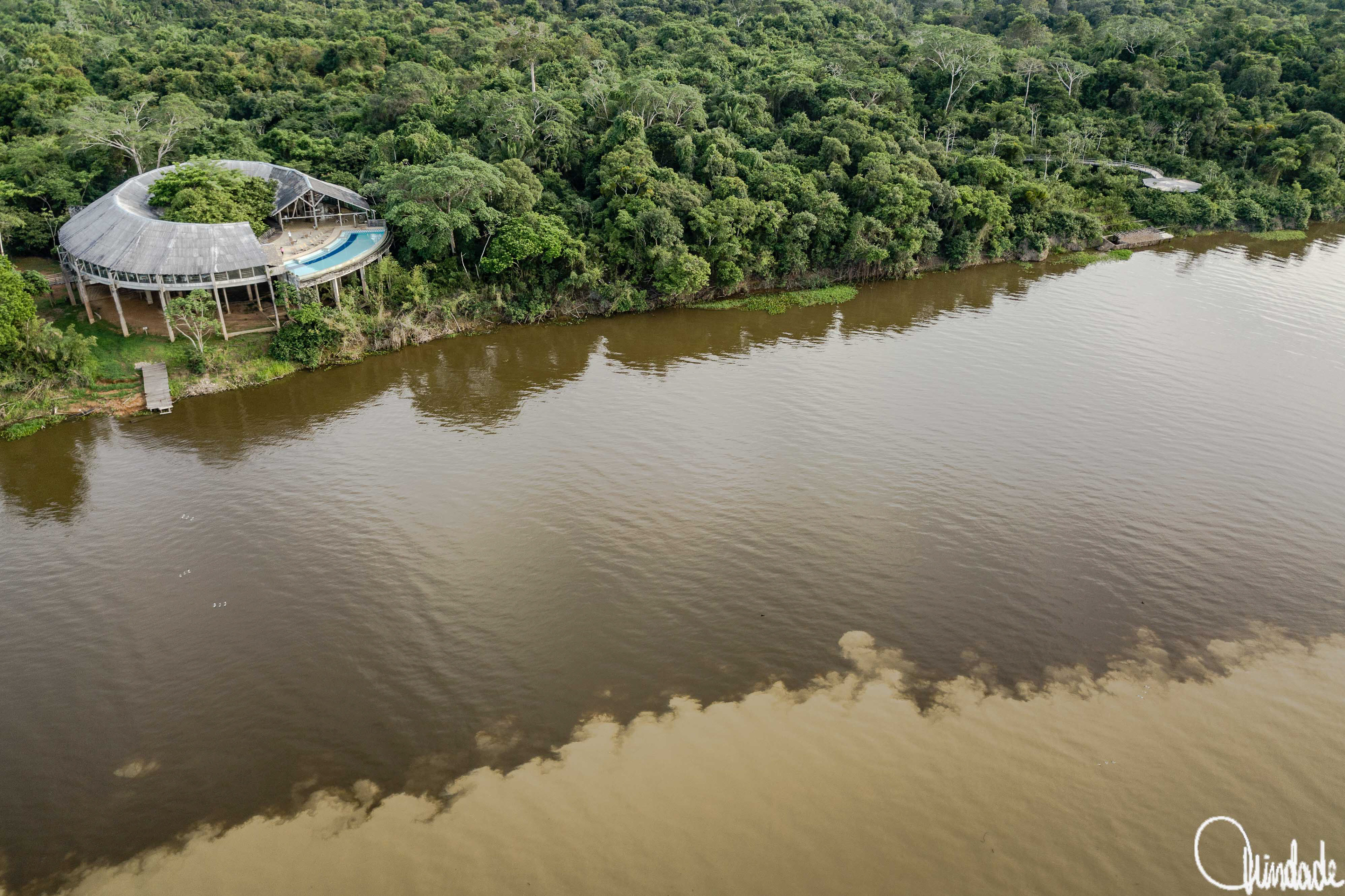 Vista aérea do Hotel Pakaas Palafitas, em Guajará-Mirim, Rondônia, mostrando sua estrutura circular elevada sobre palafitas, cercada pela floresta amazônica e situada na confluência dos rios Mamoré e Pacaas Novos. O edifício possui piscina lateral e um pequeno cais junto ao rio, destacando a integração entre arquitetura e natureza.