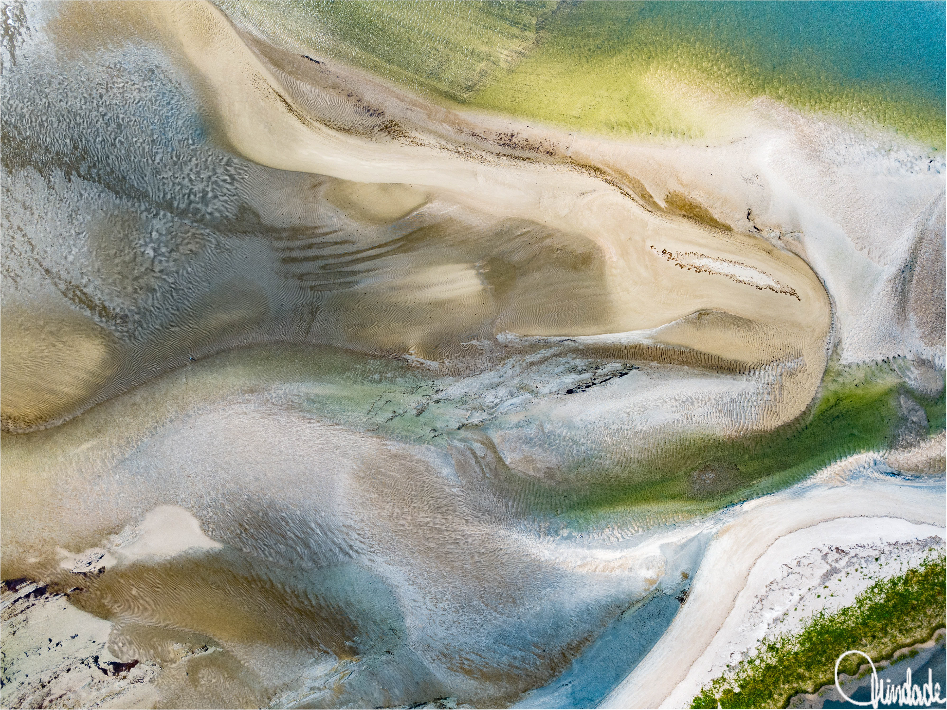 Vista aérea de estuário em Alagoas, com bancos de areia em tons bege e canais de água verde e azul. Padrões de sedimentos e curvas naturais criam composição abstrata e orgânica.
