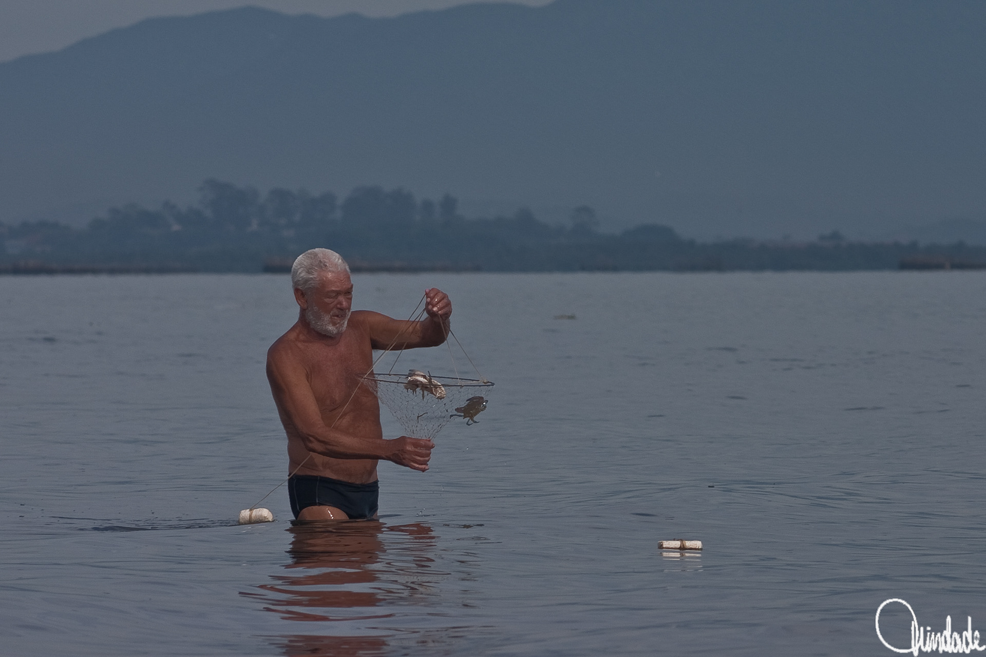 Homem em águas rasas da praia da Ilha do Governador segura rede com siris, em cena de pesca artesanal que reflete saberes rústicos e a tradição costeira carioca.