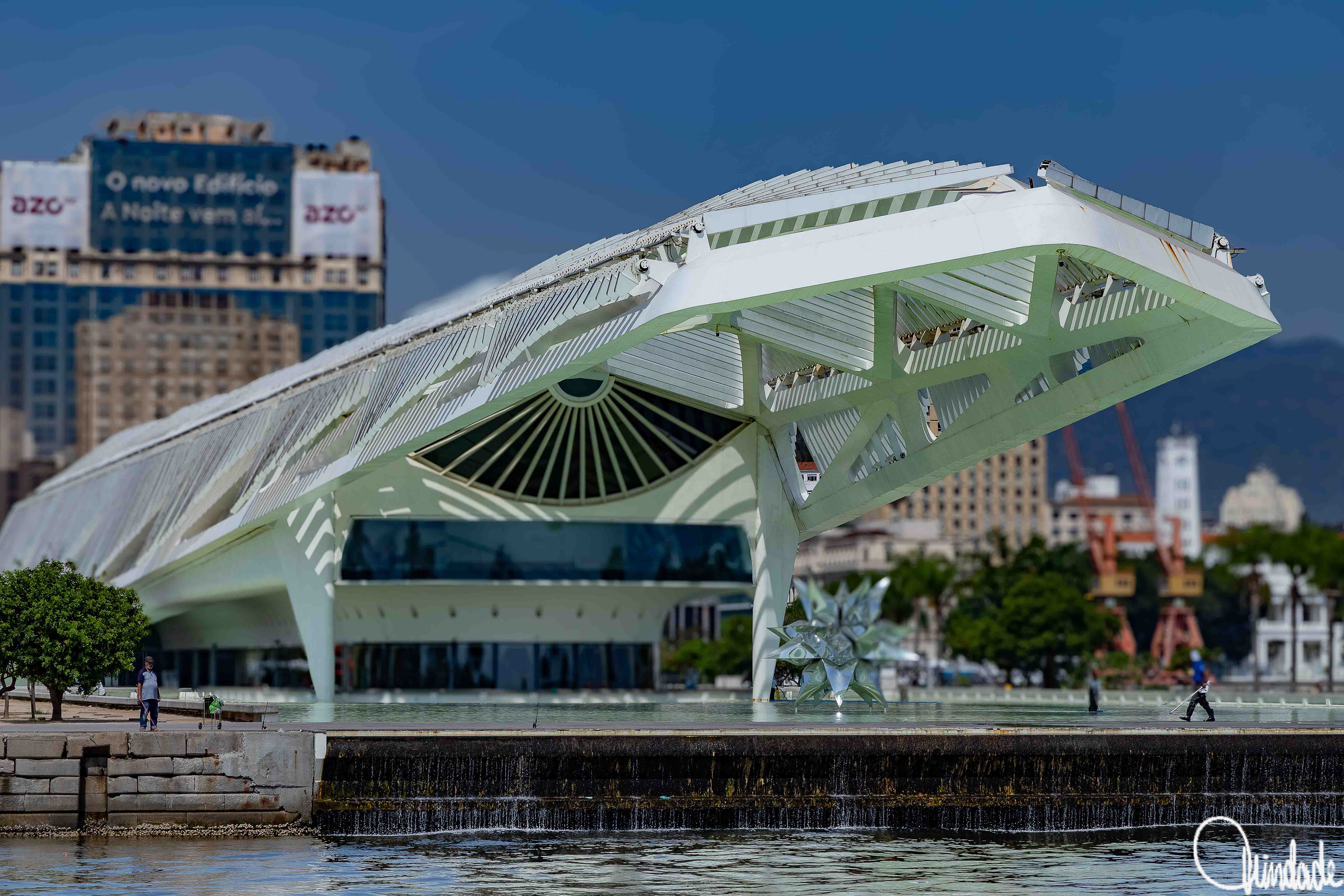 Museu do Amanhã em destaque, com sua arquitetura futurista branca refletida na água do espelho d’água, escultura metálica ao centro e prédios urbanos ao fundo (Rio de Janeiro, RJ).