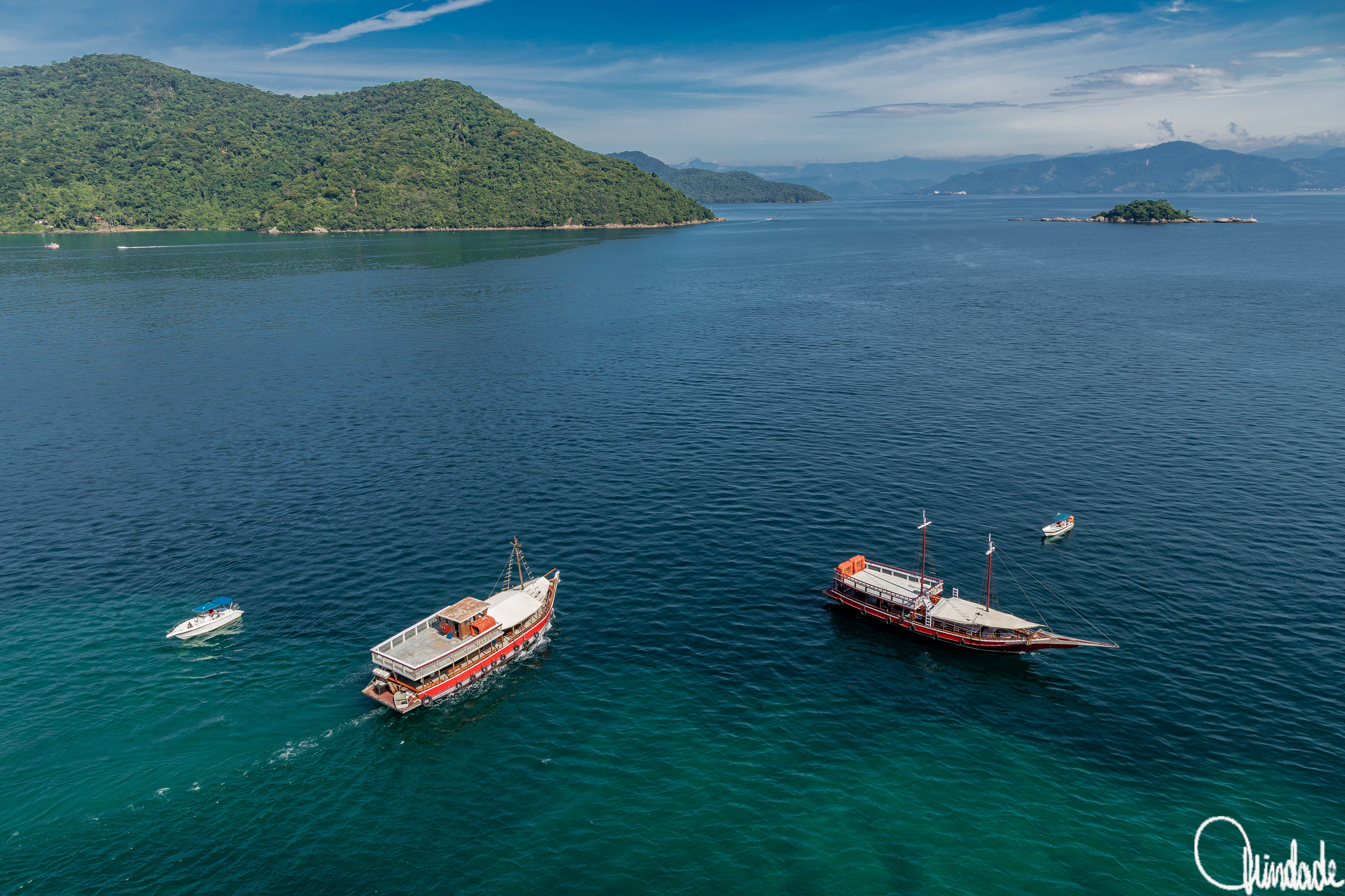 Vista costeira da Vila Abraão, Ilha Grande (RJ), com barcos no mar azul e ilhas cobertas por vegetação ao fundo.