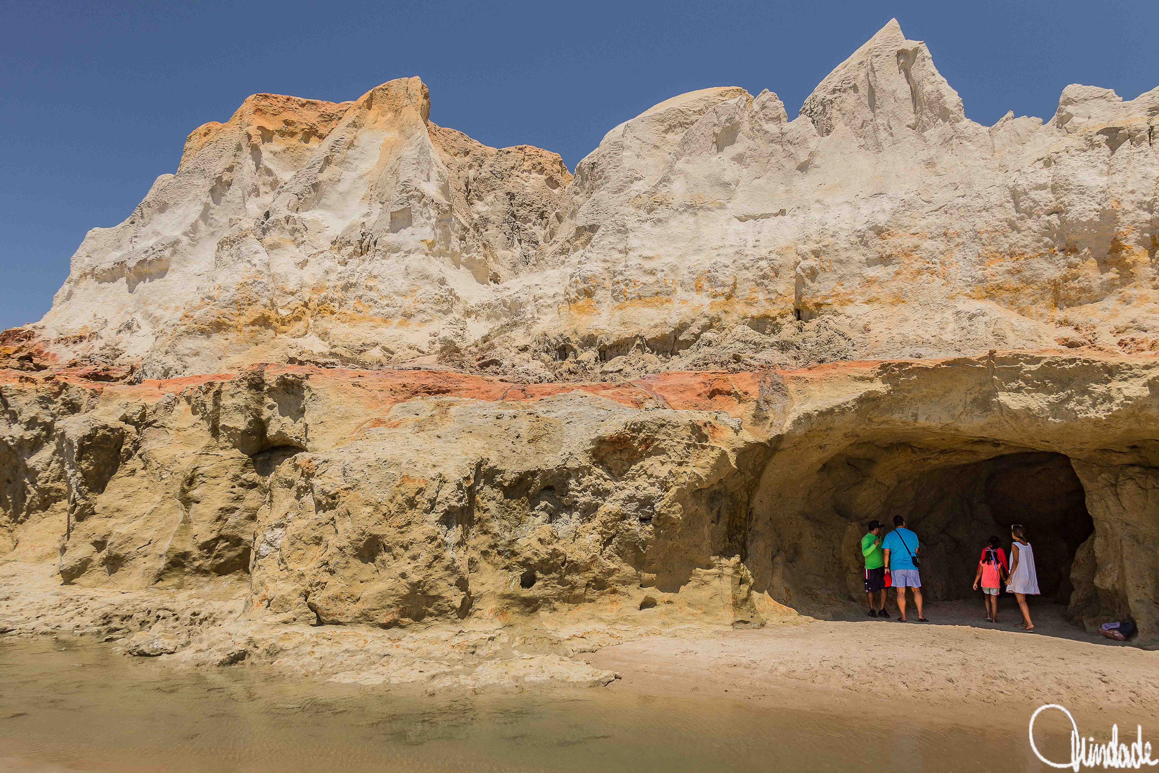 Grupo de pessoas diante de falésia multicolorida em Morro Branco, Ceará, com camadas de areia branca, amarela e avermelhada formando paredões esculpidos pela erosão, céu azul ao fundo e entrada de caverna junto à praia.
