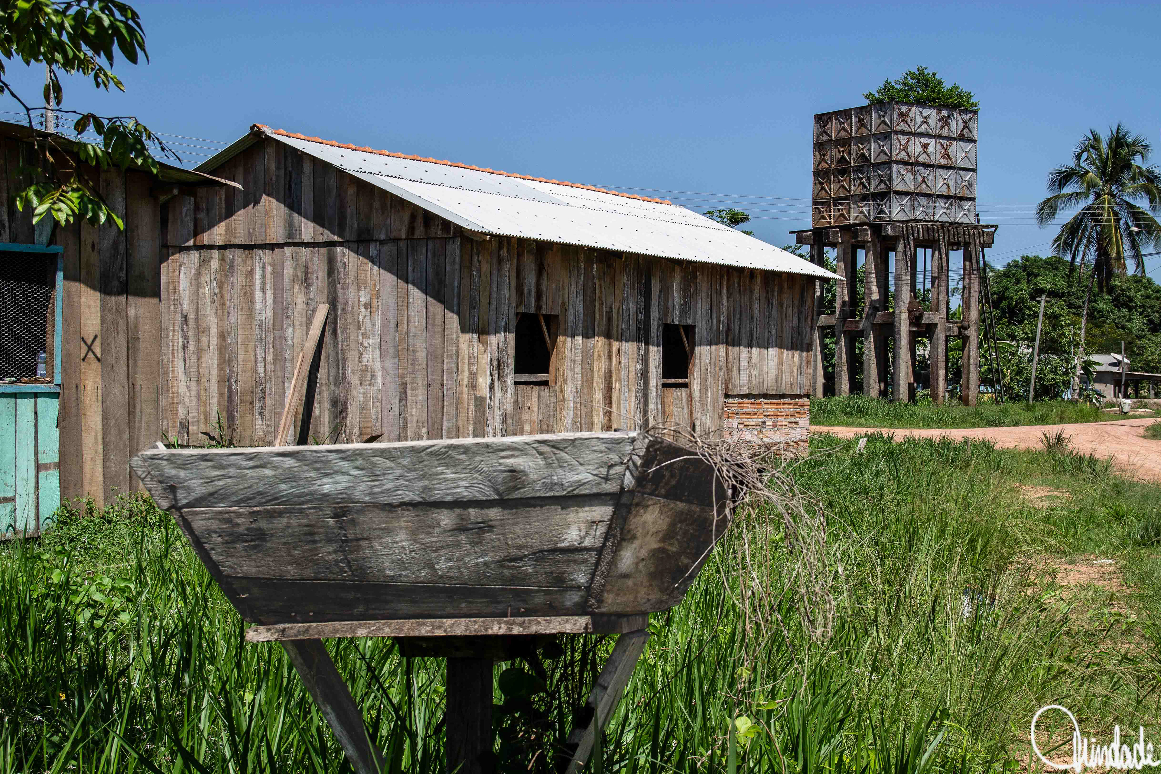 Casa rústica de madeira na Amazônia, com caixa d’água da ferrovia Madeira-Mamoré ao fundo, em Abunã, Rondônia — memória viva da história regional.