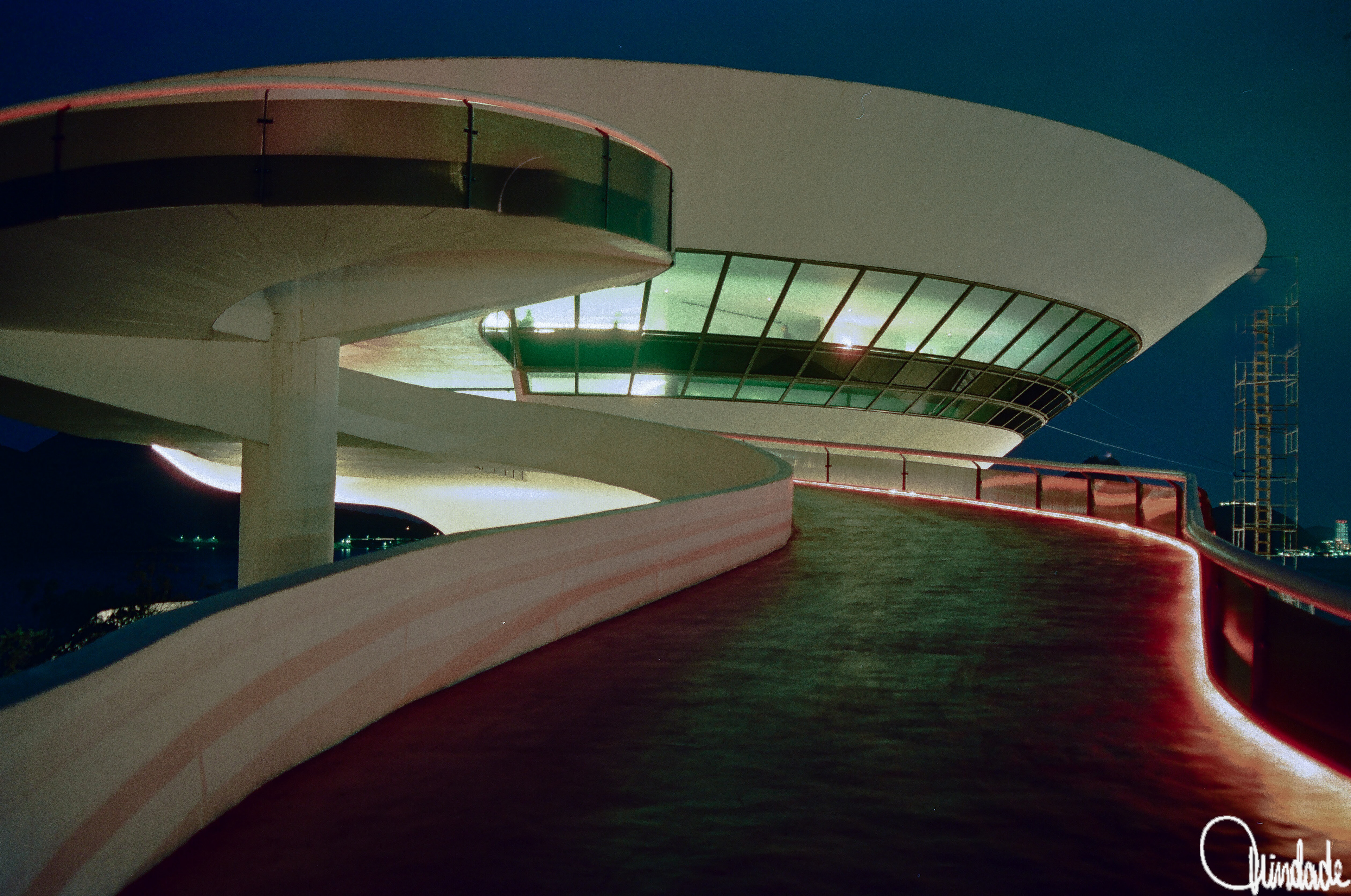 Edifício futurista em forma de disco, apoiado sobre coluna central, com rampas curvas iluminadas. Fachada de vidro reflete a luz contra o céu noturno, destacando linhas orgânicas da arquitetura modernista.