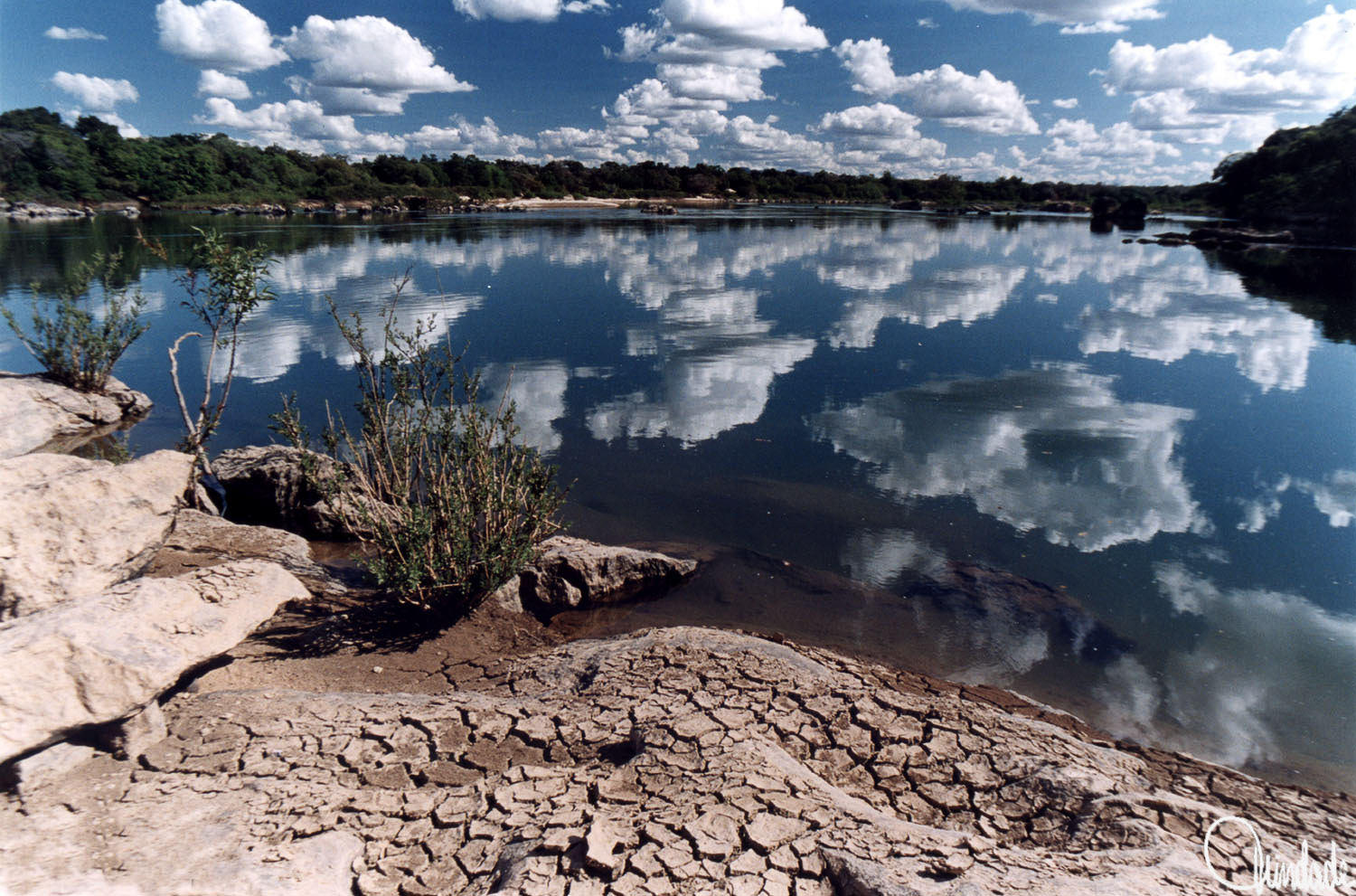 Rio Paranã em Tocantins com reflexo das nuvens no céu azul sobre a superfície calma da água, margem seca com rachaduras e vegetação esparsa em primeiro plano, e linha de árvores verdes ao fundo.