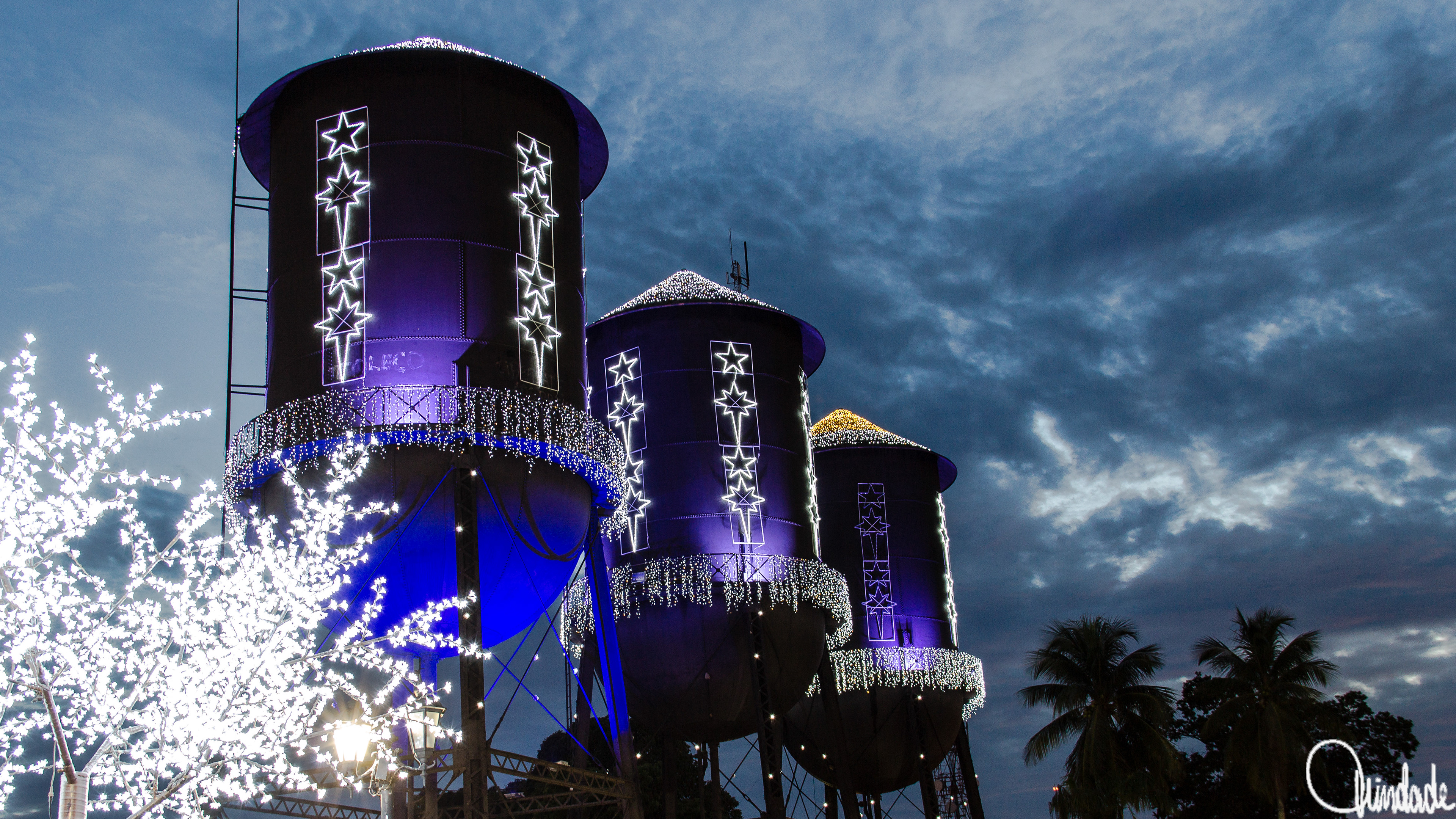 Três grandes torres cilíndricas de água iluminadas em azul e roxo, com padrões de estrelas brilhantes e luzes decorativas. Uma árvore coberta de luzes brancas aparece em primeiro plano sob céu nublado ao entardecer.