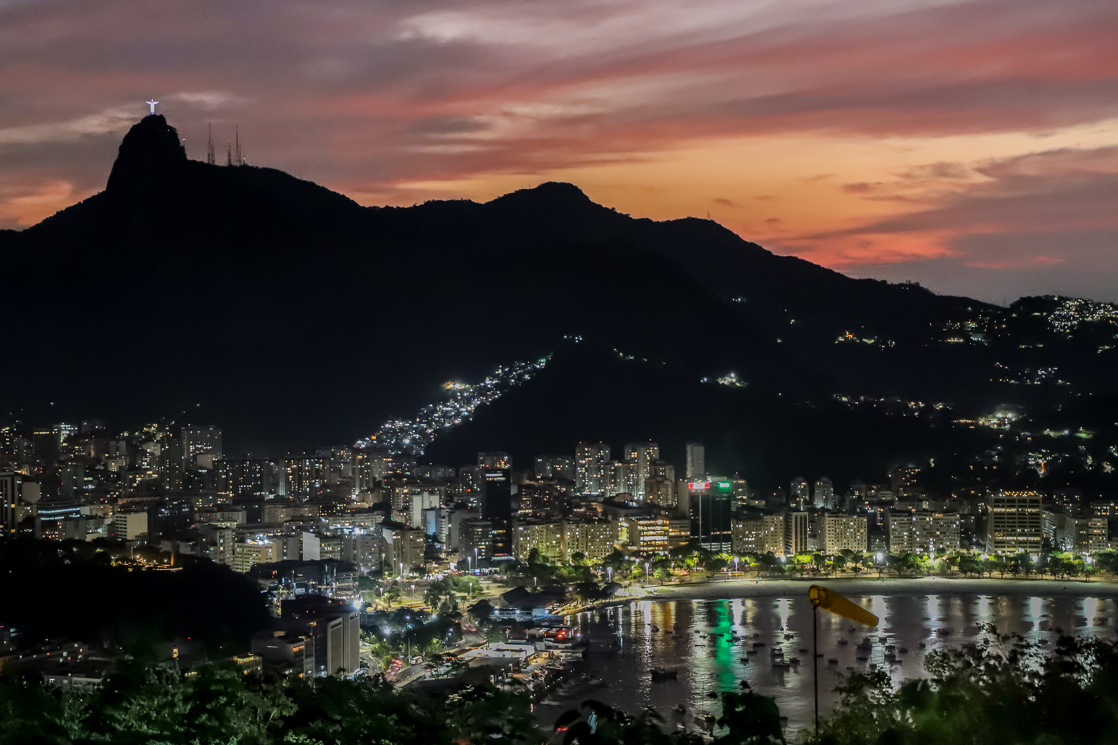    "Vista noturna da enseada de Botafogo no Rio de Janeiro com reflexo das luzes da cidade na água, serra da Carioca ao fundo e Cristo Redentor iluminado no alto do Corcovado sob céu colorido ao pôr do sol.