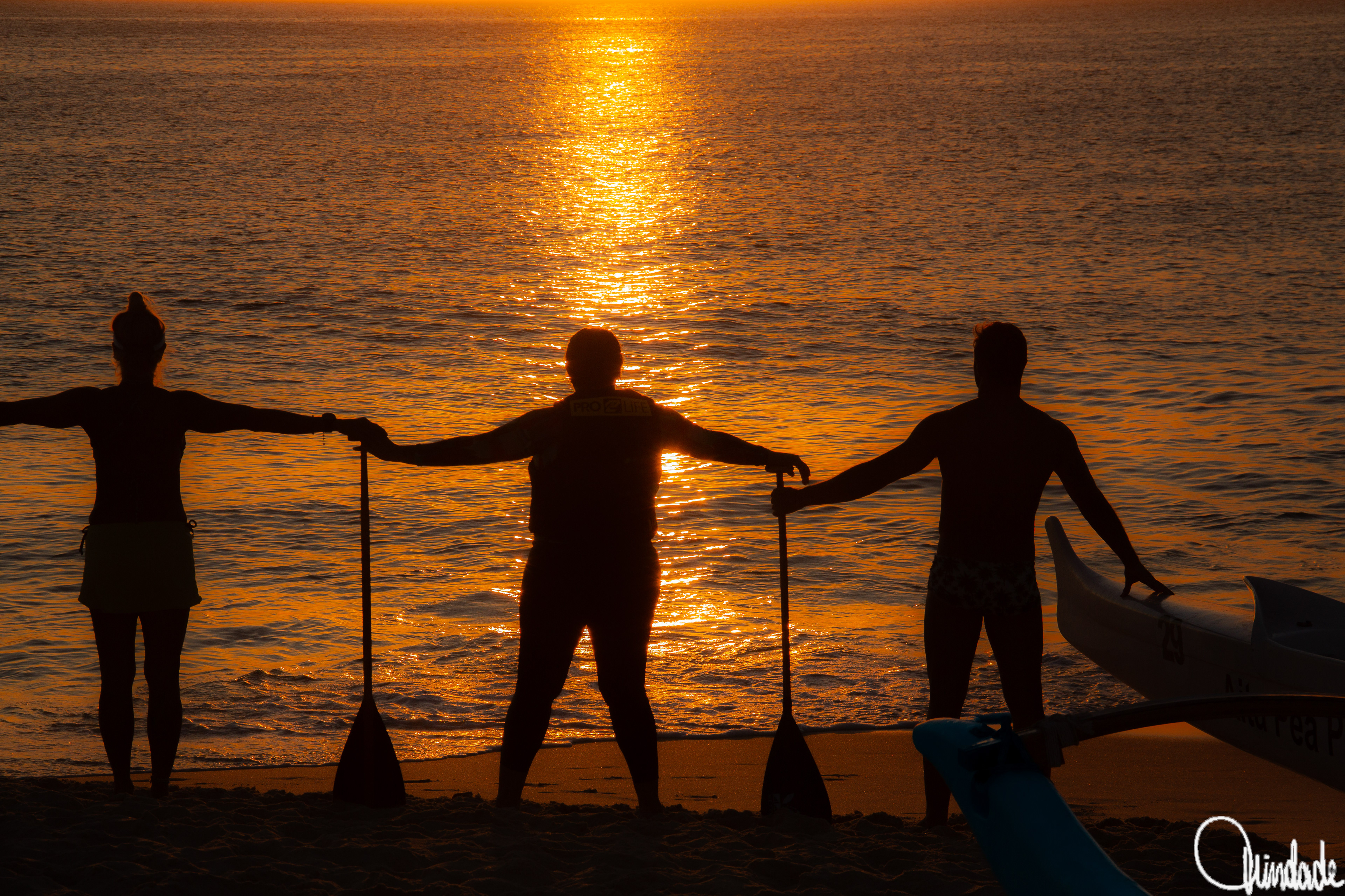Três praticantes de canoagem em silhueta ao pôr do sol na praia de Camboinhas, com remos erguidos e canoa à beira-mar, em cena rústica e coletiva de navegação.