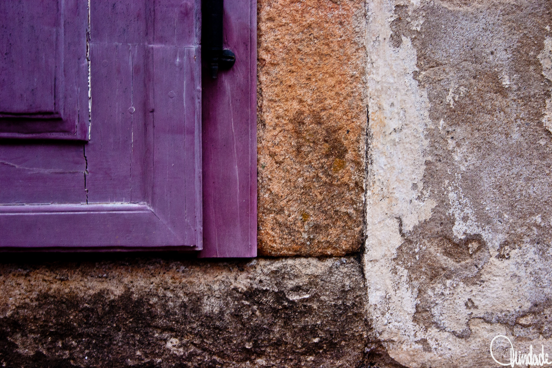 Close de portal de casa colonial com parede de pedra e reboco desgastado. Porta ou janela de madeira pintada em roxo profundo, com ferragem preta, contrasta com tons terrosos da parede.