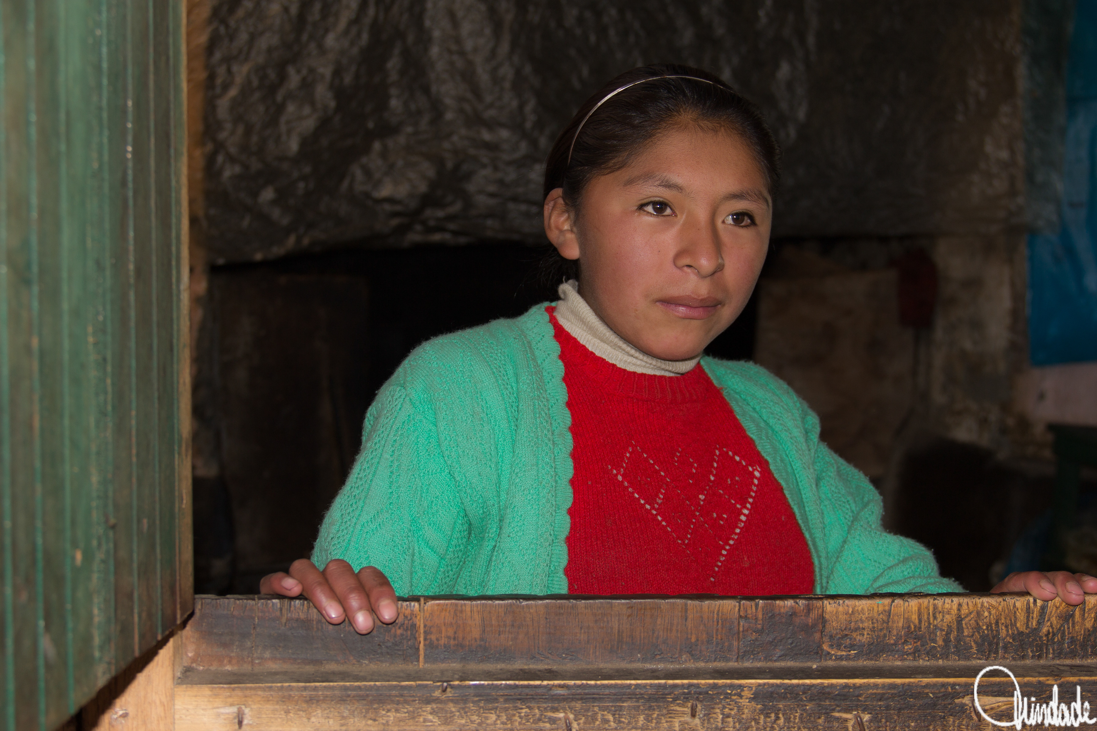 Jovem com suéter vermelho e cardigã verde, em ambiente rústico, iluminada por luz suave, representando moradora da Cordilheira dos Andes no Peru.