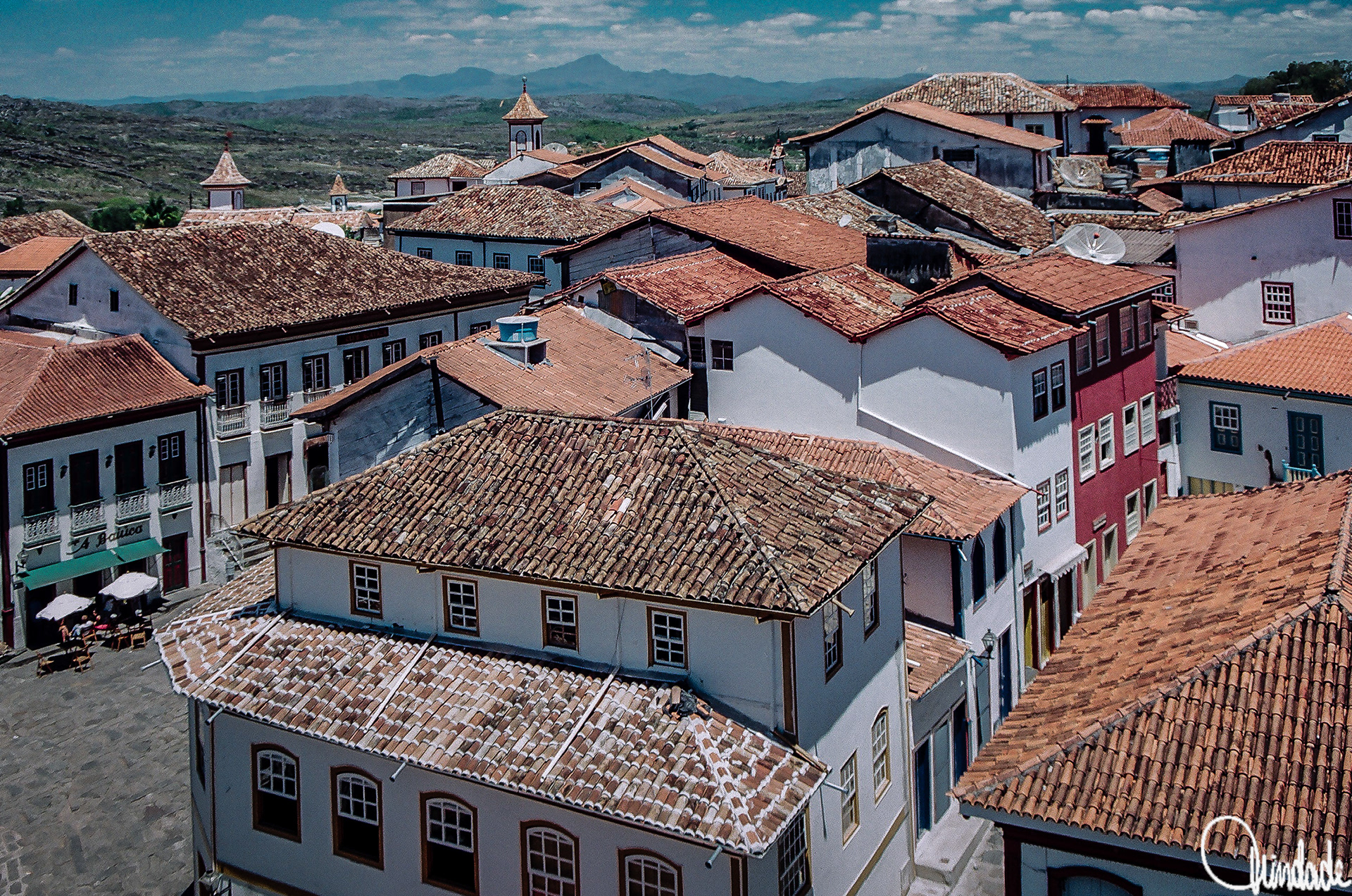 Vista panorâmica de cidade histórica com casas coloniais de paredes brancas, janelas e sacadas de madeira, telhados de telha vermelha. Ao fundo, colinas e montanhas sob céu parcialmente nublado.