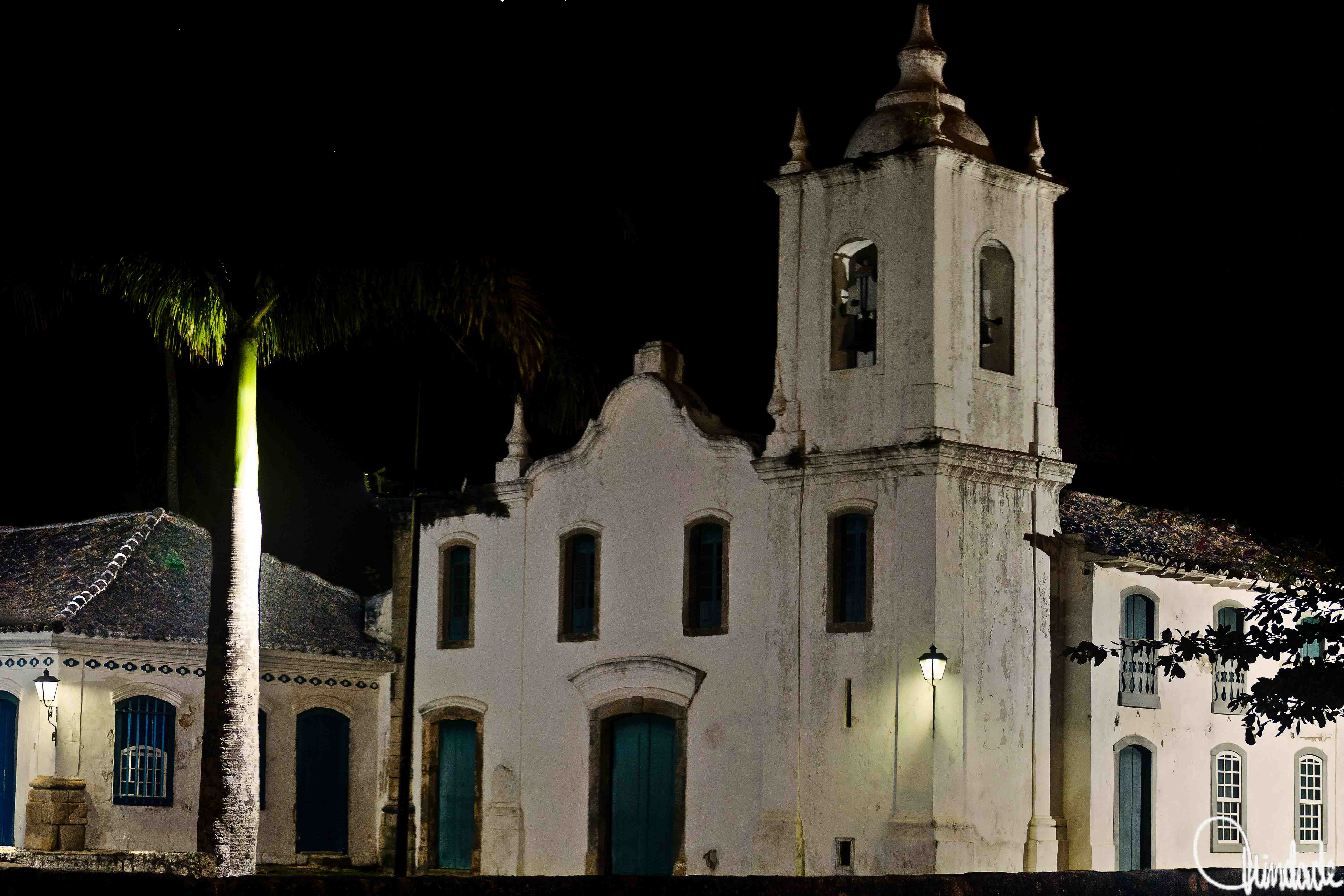 Igreja de Santa Rita iluminada à noite no centro histórico de Paraty, Rio de Janeiro, com fachada branca envelhecida, portas e janelas em azul-esverdeado e torre com cúpula e sino.