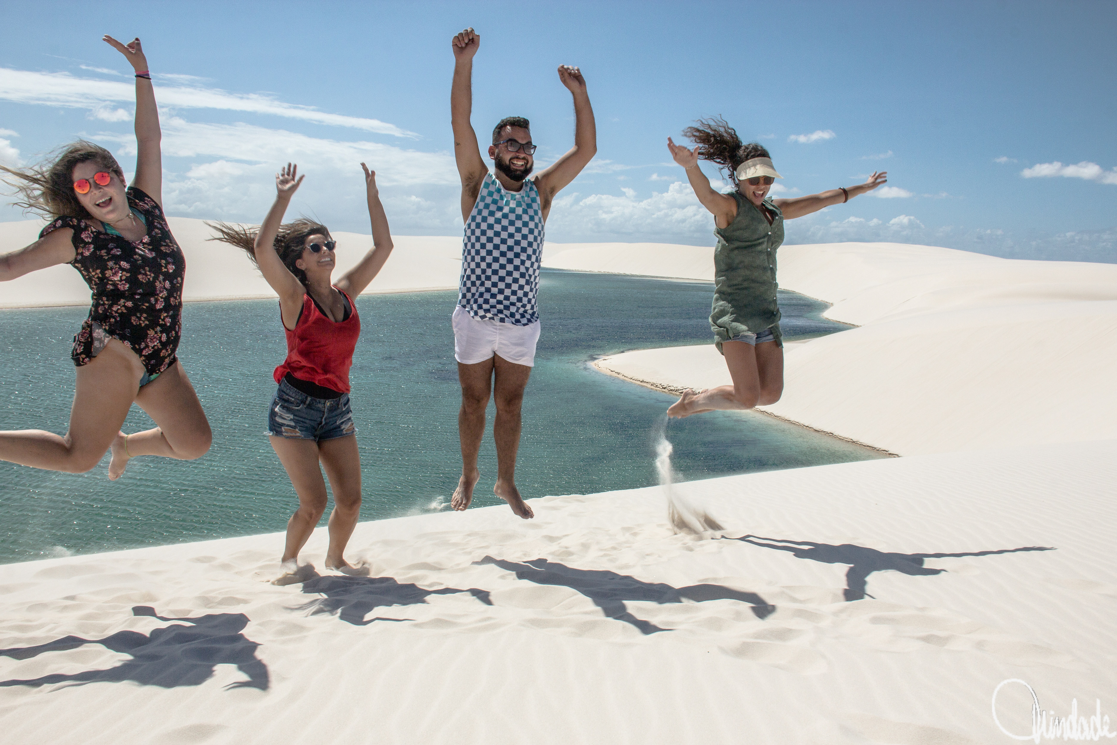 Quatro pessoas saltando sobre dunas brancas diante de lagoas azul-turquesa nos Lençóis Maranhenses, em cena vibrante de alegria e movimento na paisagem natural.