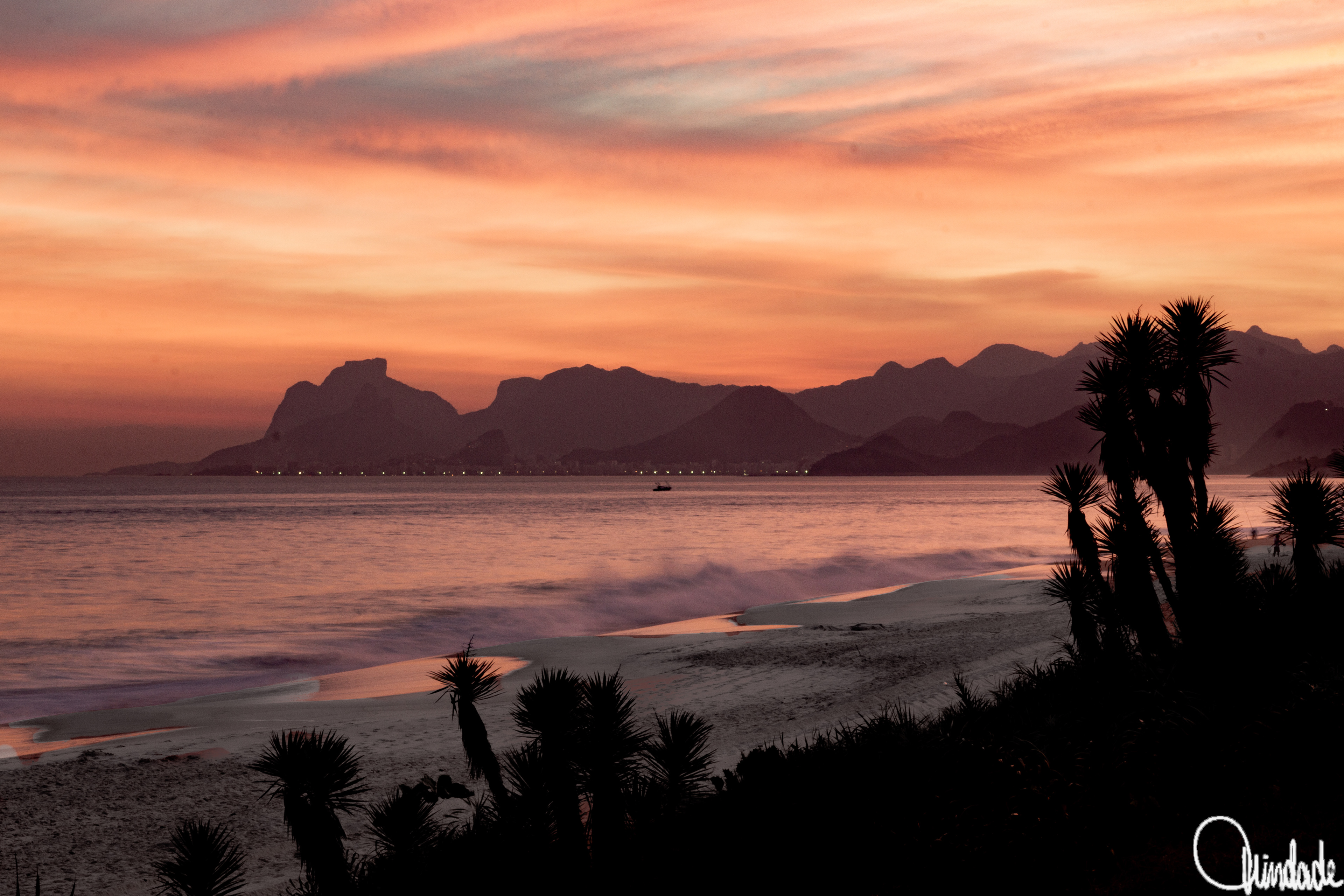 Praia de Camboinhas em Niterói, Rio de Janeiro, com vegetação costeira em primeiro plano, mar refletindo tons alaranjados do pôr do sol e montanhas cariocas ao fundo parcialmente cobertas por névoa.